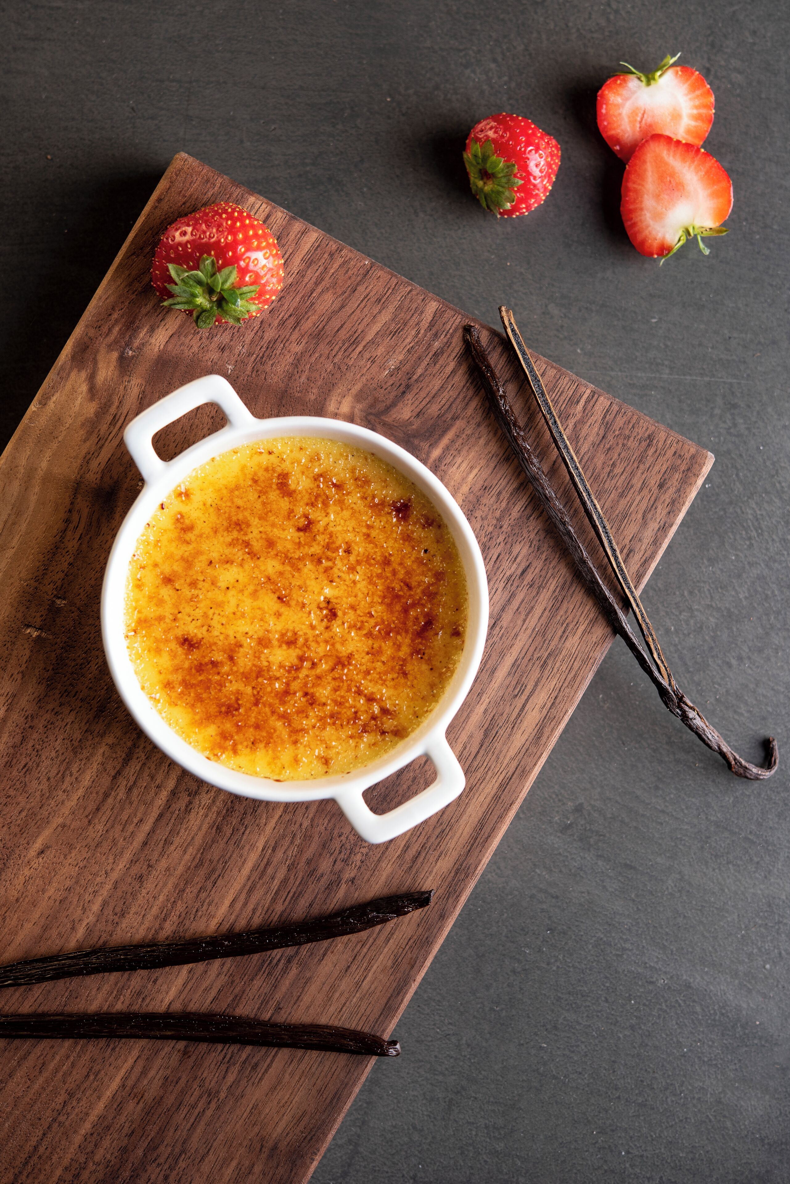 En skål creme brûlée på en træplade med vaniljestænger og jordbær.