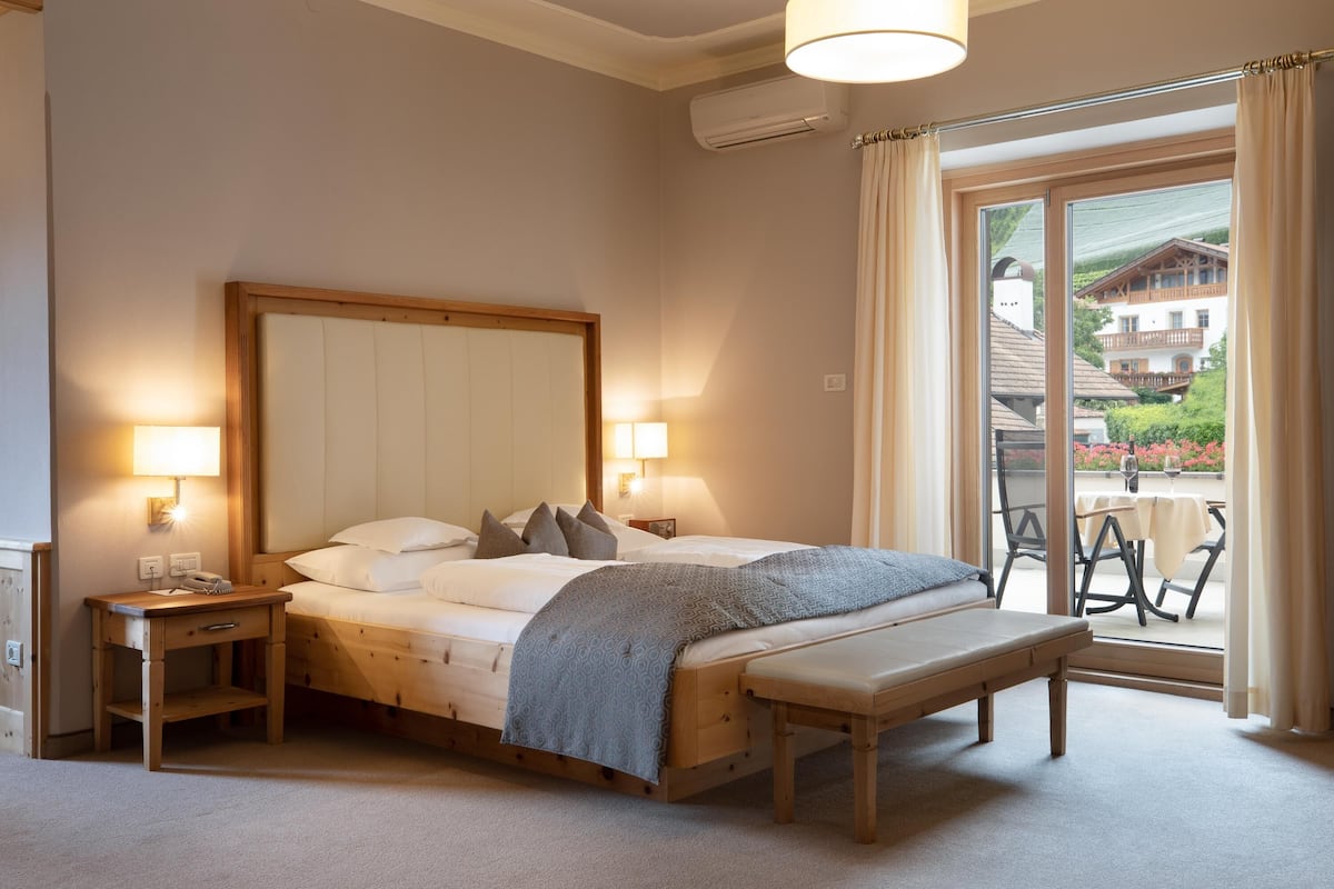 A bedroom with a large bed, bedside tables, a bench, and a view of a balcony with outdoor furniture.