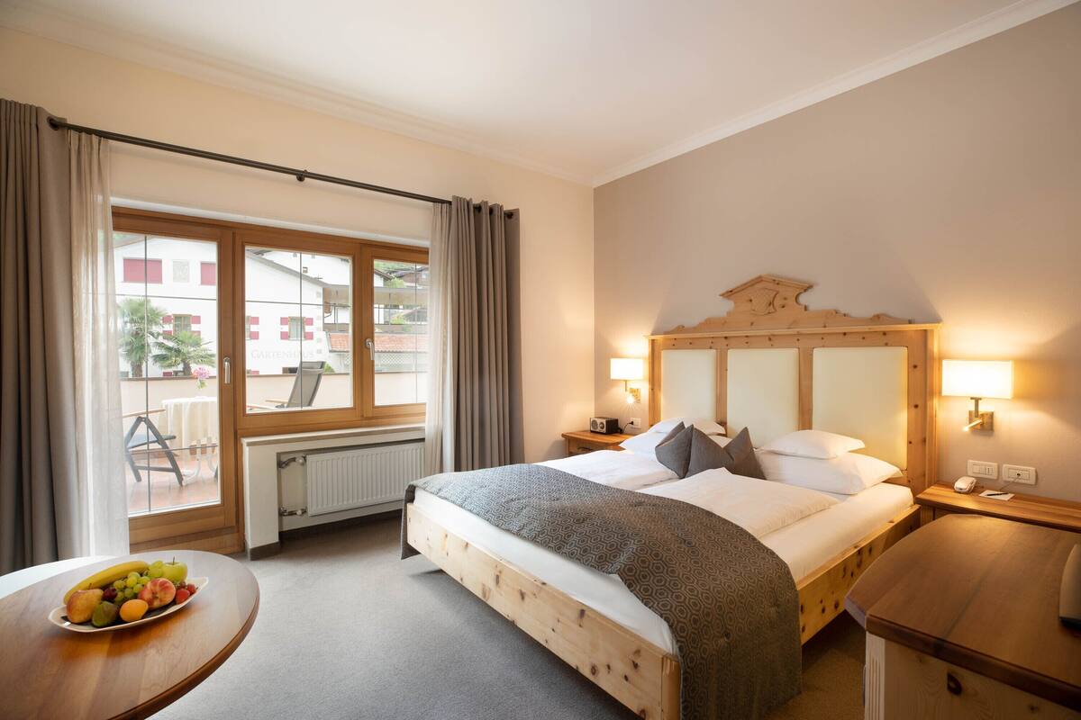 A hotel room with a bed, bedside table, lamp, and a view of the outside.