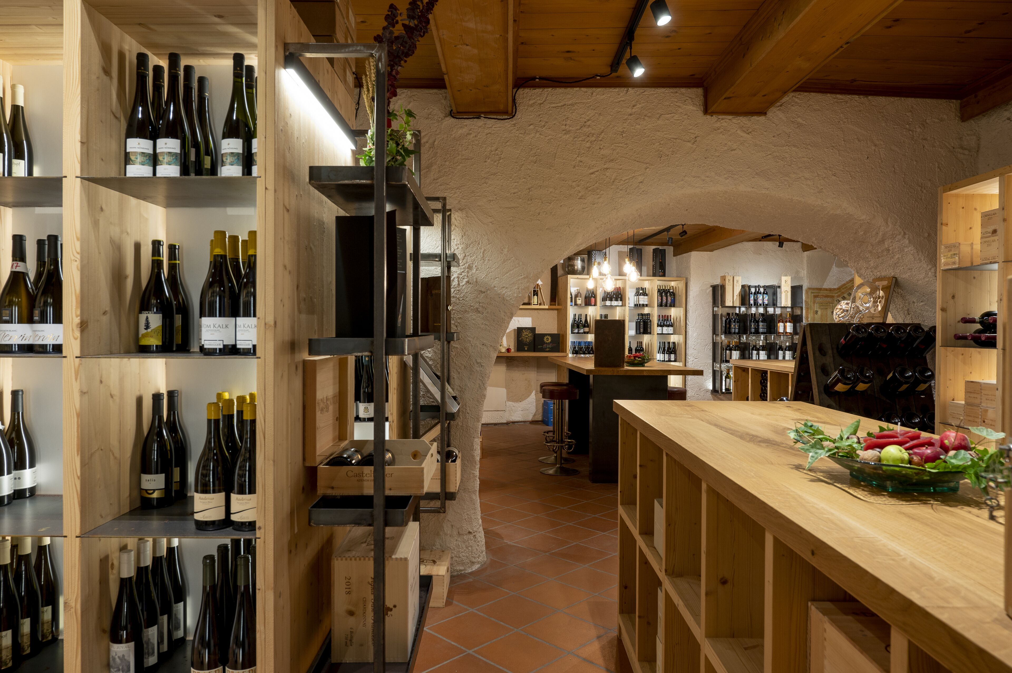 A wine cellar with wooden shelves and racks, a wooden counter with a bowl of fruit, and a rustic stone wall.