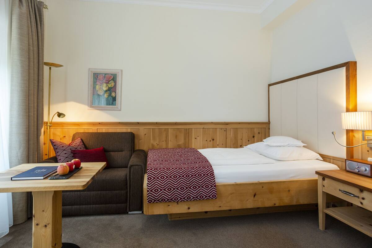 A room with a bed, a sofa, a table with apples and a book, a lamp, and a painting on the wall.
