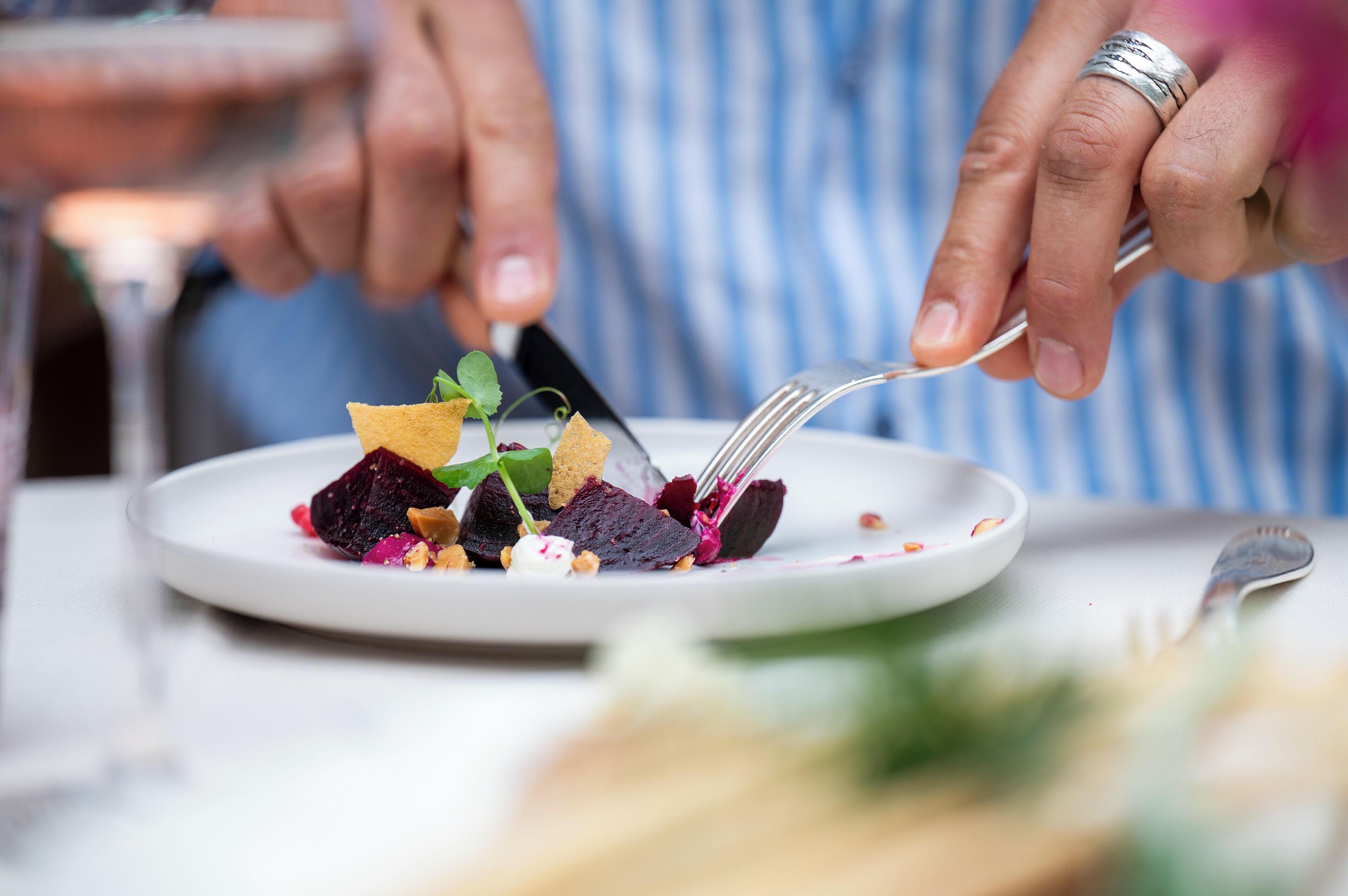 A person holding a fork with a plate of food, including beets and greens, on a table.