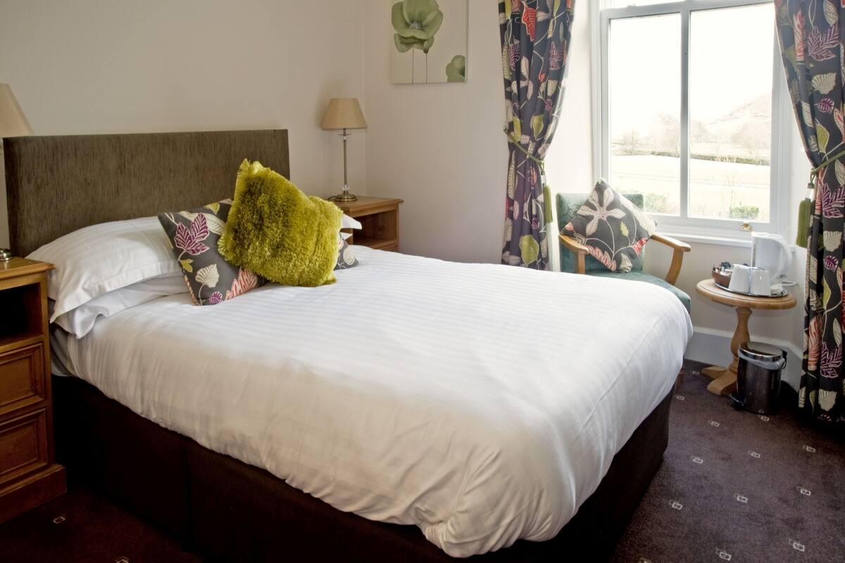 A neatly made bed with white linens and a green throw pillow, a wooden nightstand with a lamp, a chair with a floral cushion, and a small table with a coffee pot.
