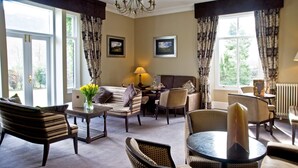 A well-lit living room with a sofa, armchairs, a coffee table, and a dining table. There are two windows with curtains, a chandelier, and framed pictures on the walls. - New Dungeon Ghyll Hotel (Ambleside)
