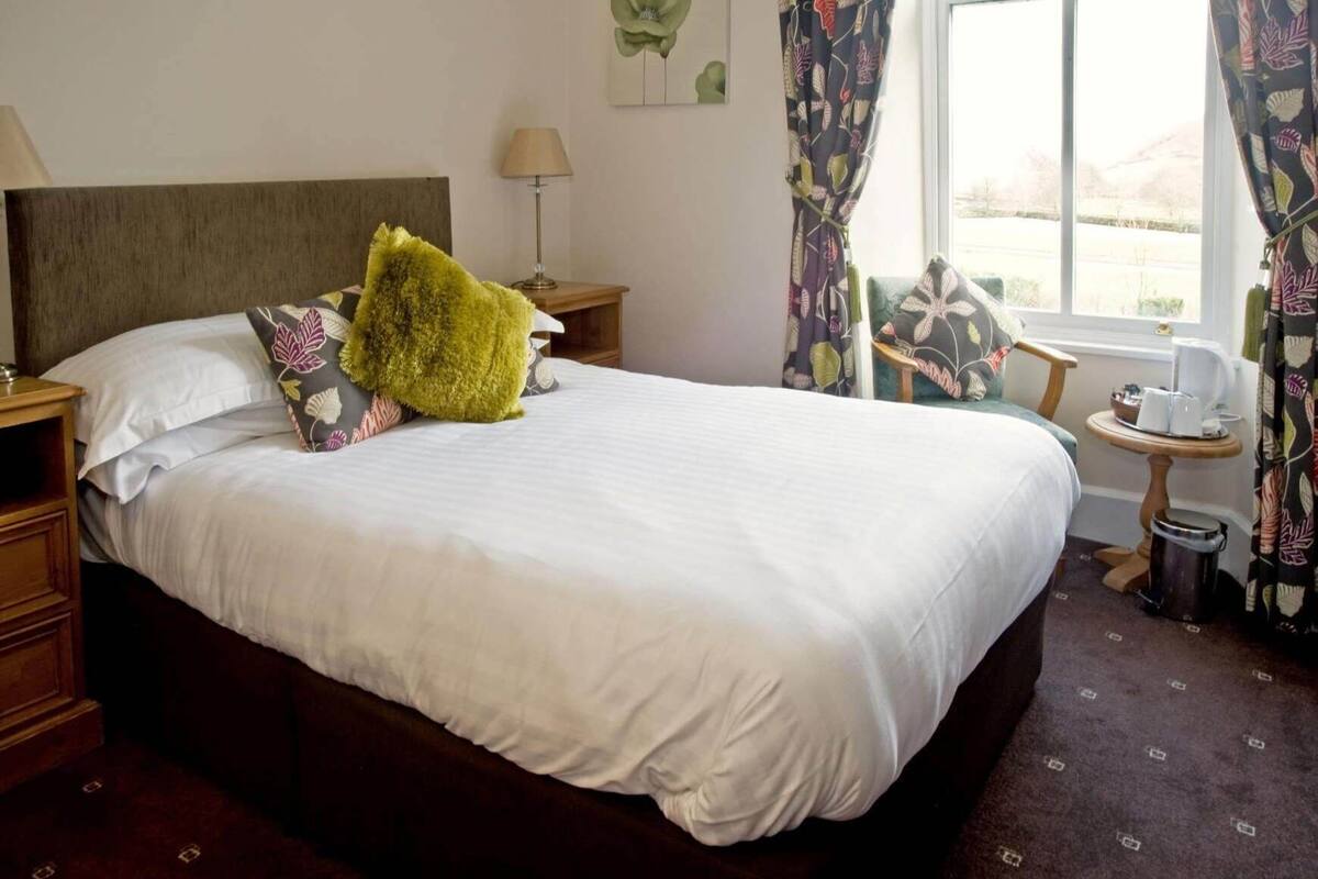 A hotel room with a large bed, a nightstand, a lamp, a small table with a kettle, and curtains.
