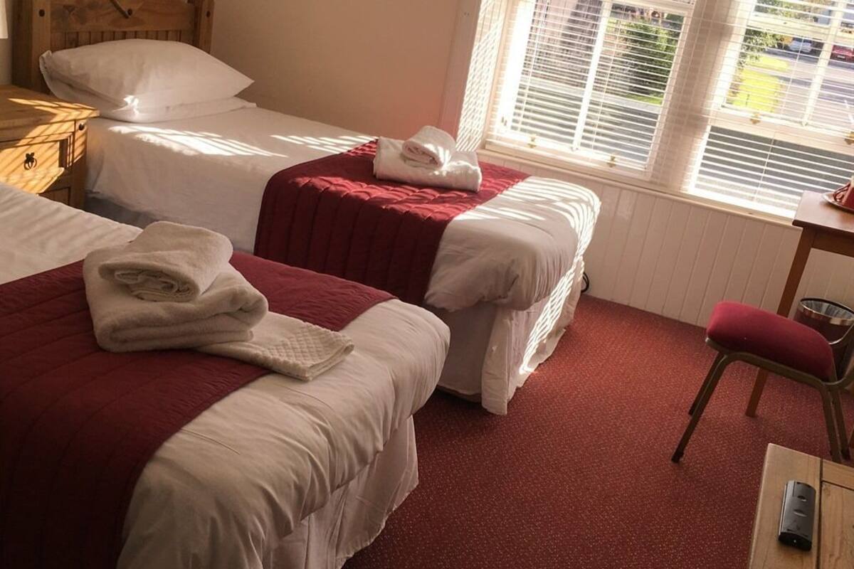 Quarto de hotel com duas camas de solteiro, uma mesinha de cabeceira de madeira, uma escrivaninha com cadeira e uma janela com persianas.