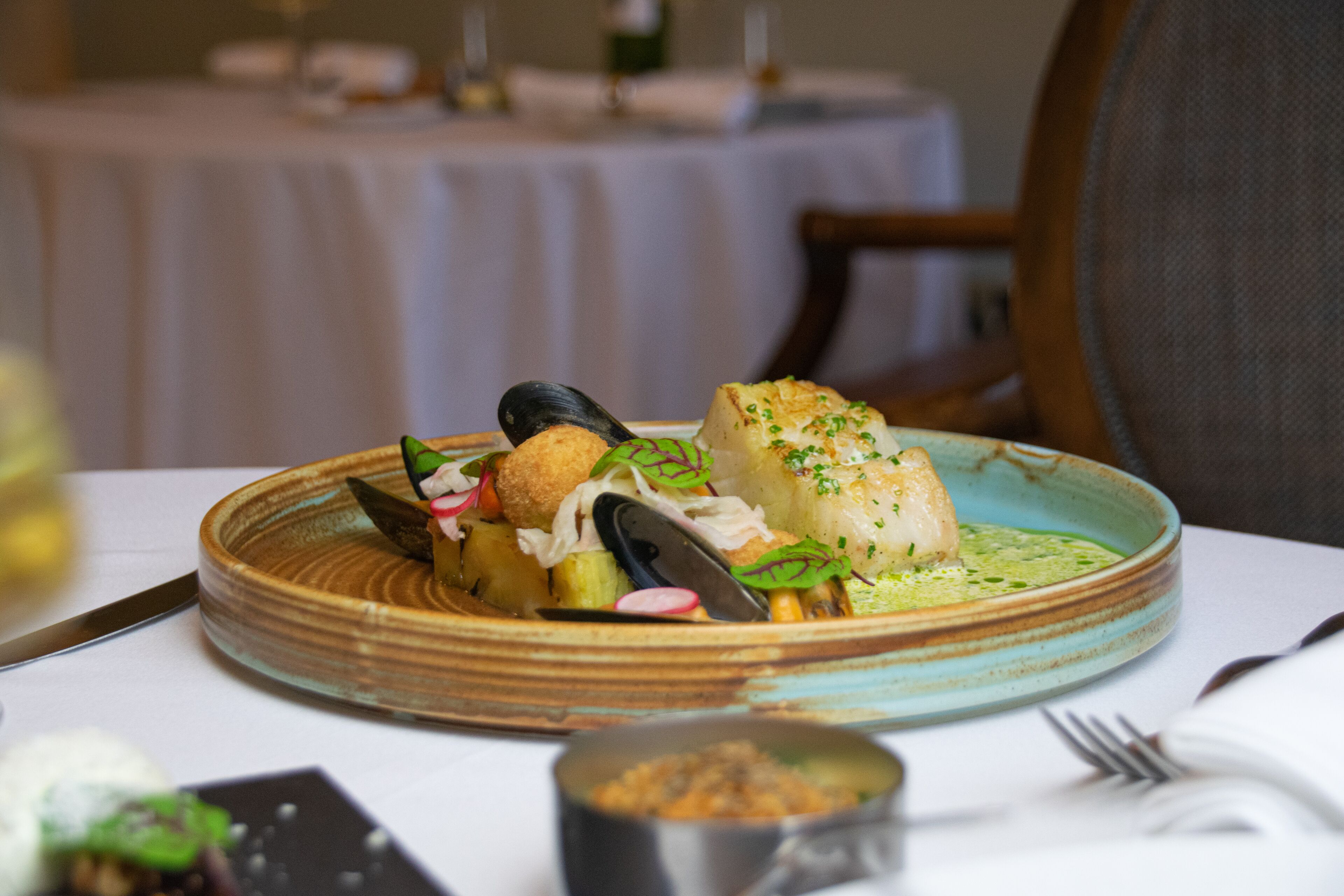 A plate of seafood with mussels, a piece of fish, and a breaded item, served on a ceramic plate with a green sauce.