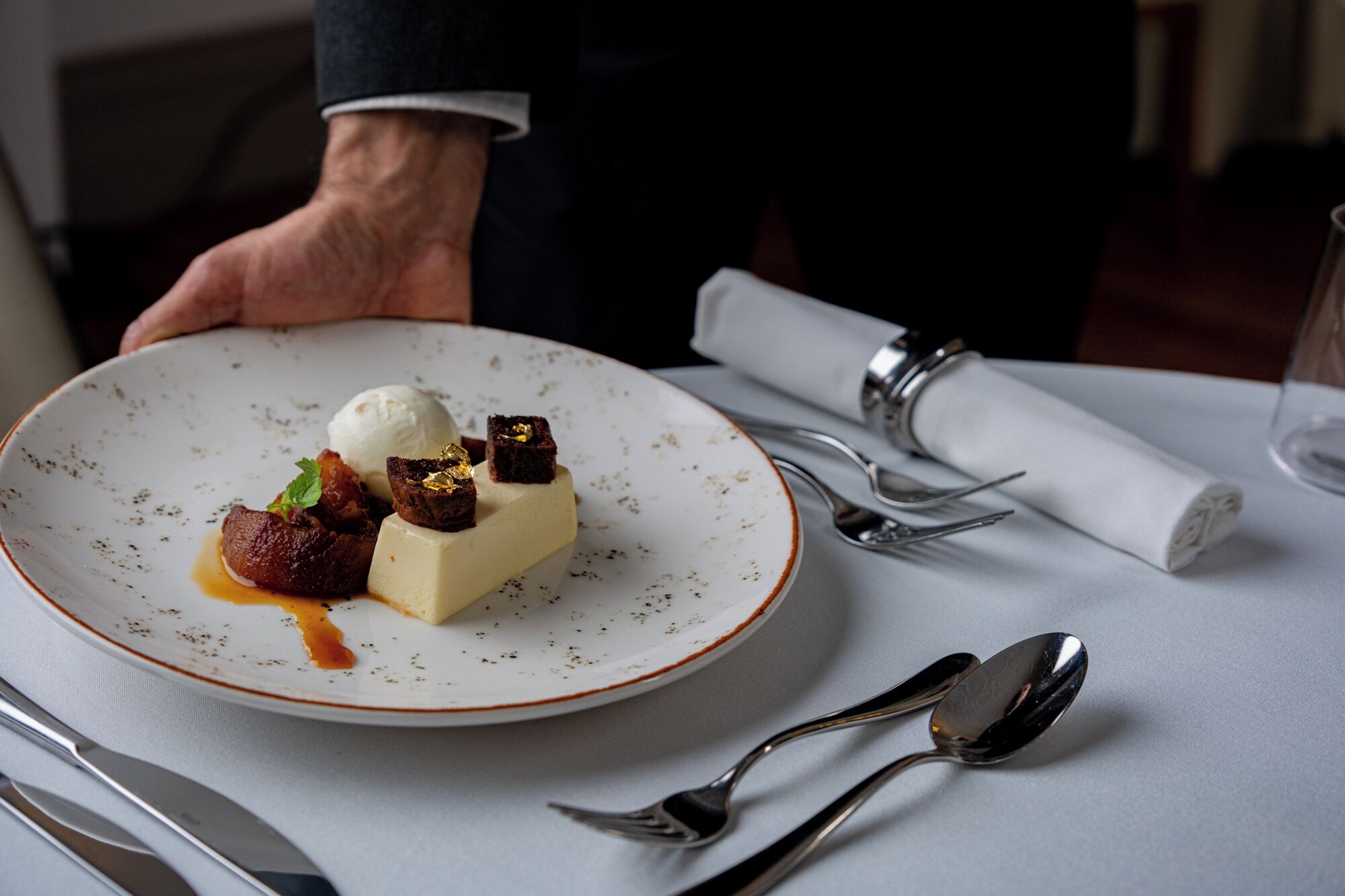 Un piatto dessert con una pallina di gelato, un cubetto che sembra formaggio e un pezzo di frutta, accompagnati da un cucchiaio e una forchetta su un tavolo.