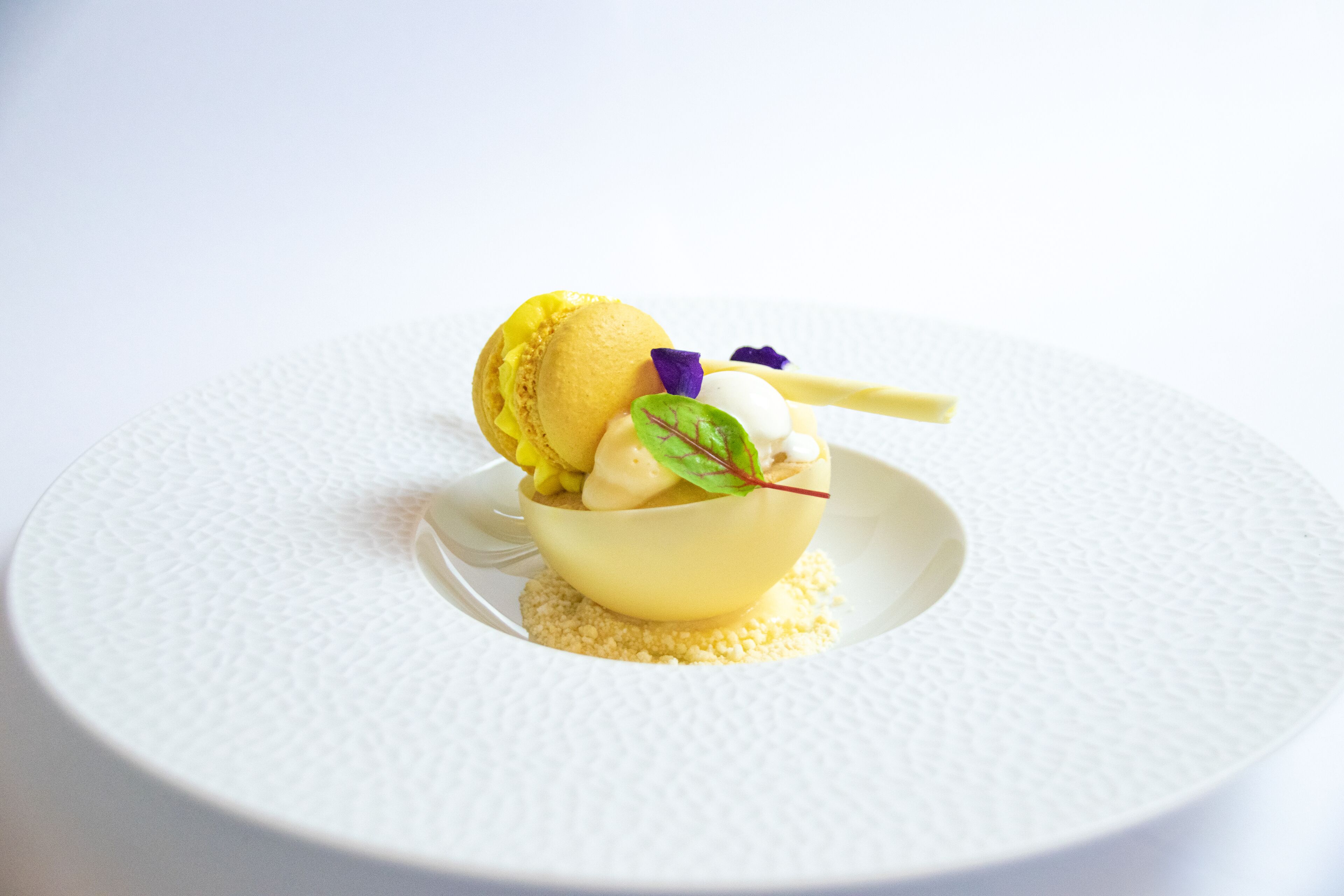 A dessert on a white textured plate with a yellow bowl, a scoop of ice cream, and a mint leaf garnish.