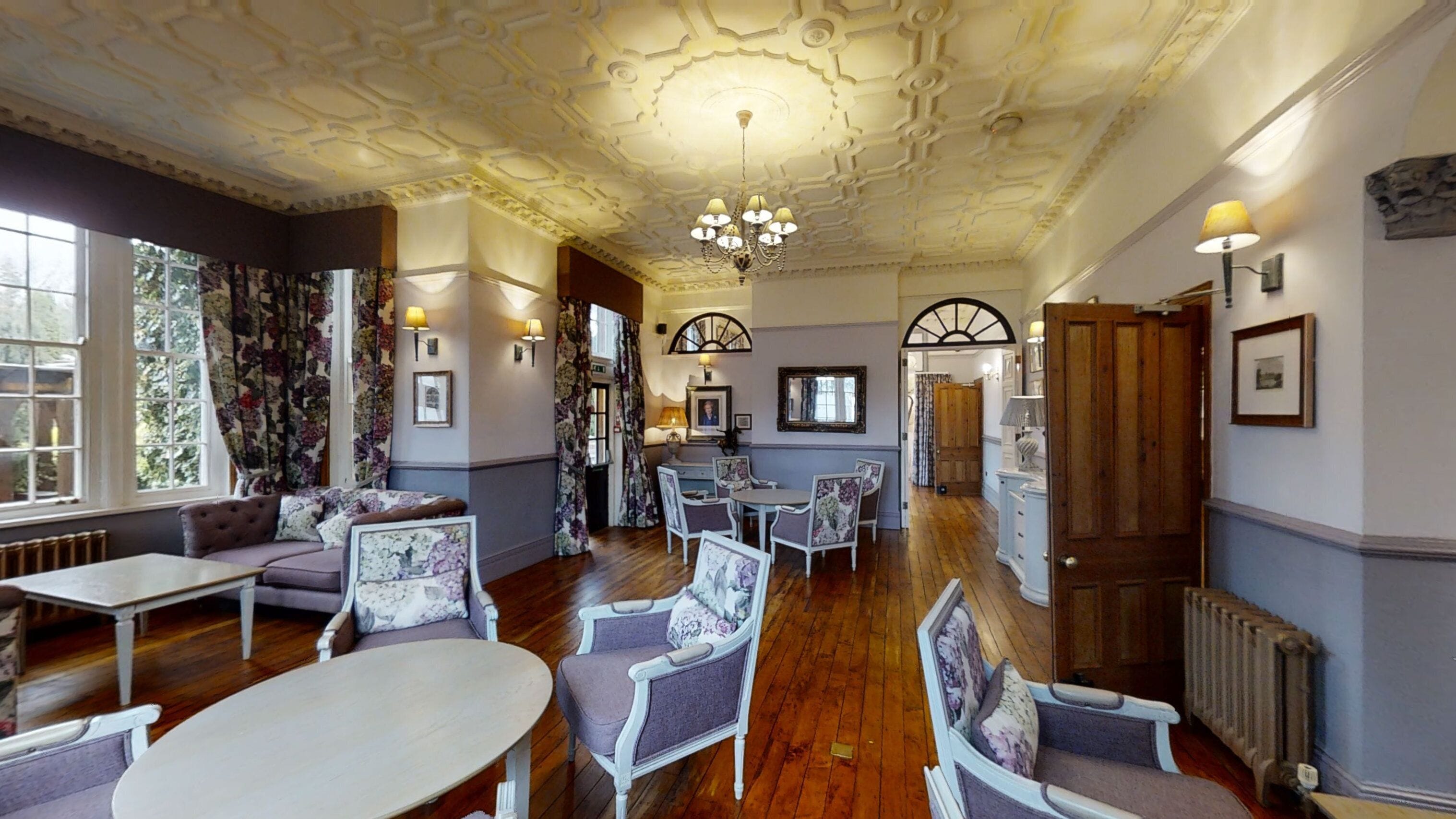A room with a wooden floor, white furniture, and a chandelier. There are two windows with floral curtains and a door leading to another room.