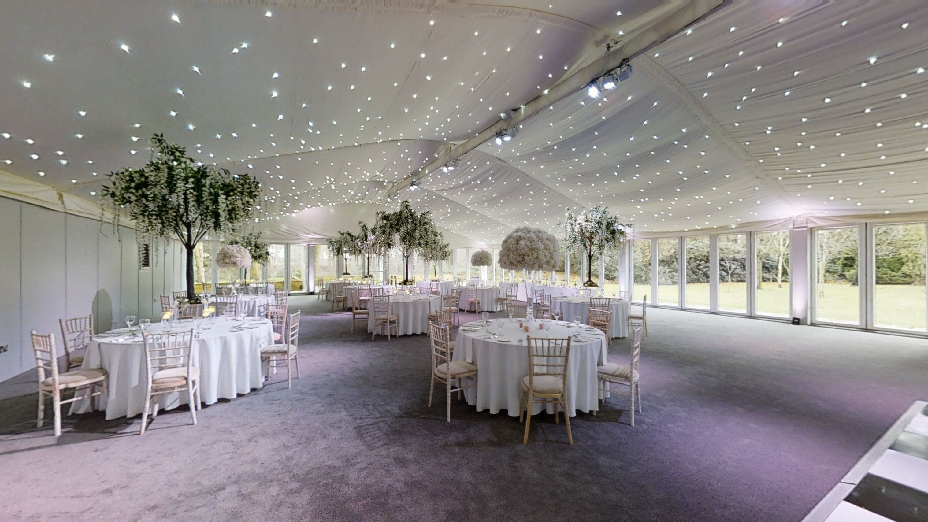 Een evenementenruimte met tenten, ronde tafels en stoelen, grote ramen en hangende bloemdecoraties.