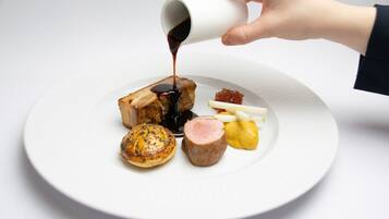 A white plate with various food items, including a piece of meat and a pastry, being drizzled with sauce.