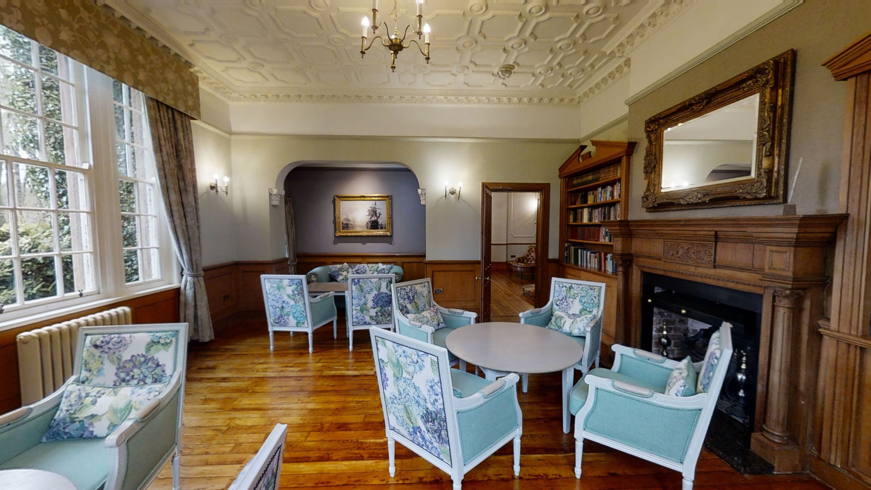 A room with a wooden floor, a round table, floral-patterned chairs, a fireplace, and a bookshelf.