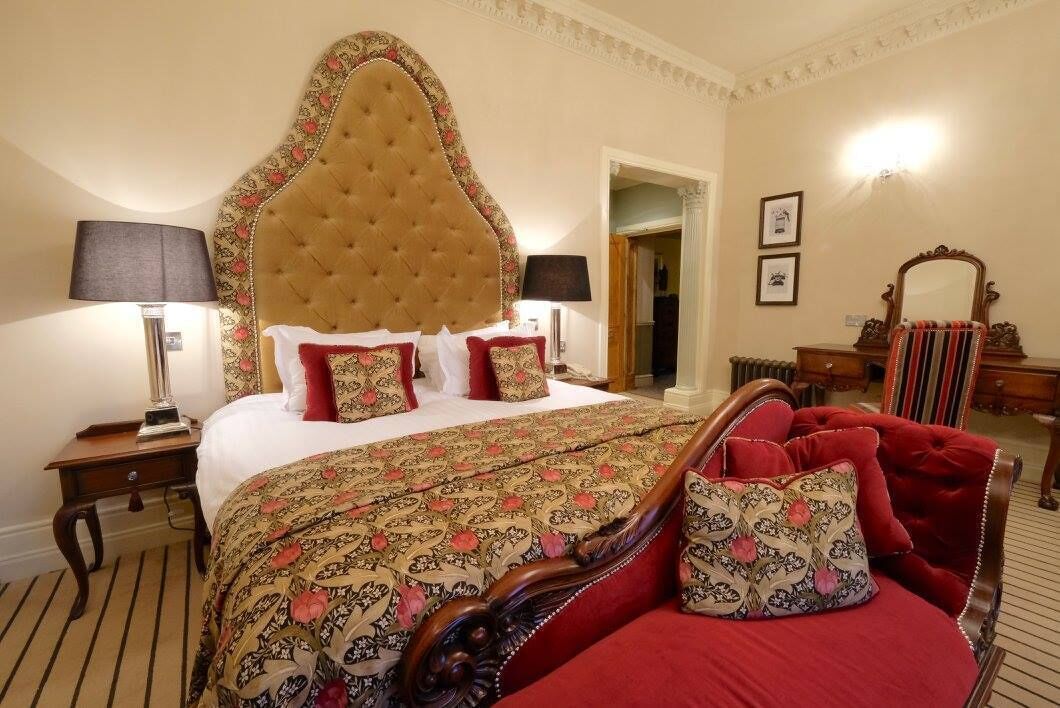 A hotel room with a large bed, a red upholstered chair, a wooden nightstand, a lamp, and a mirror.