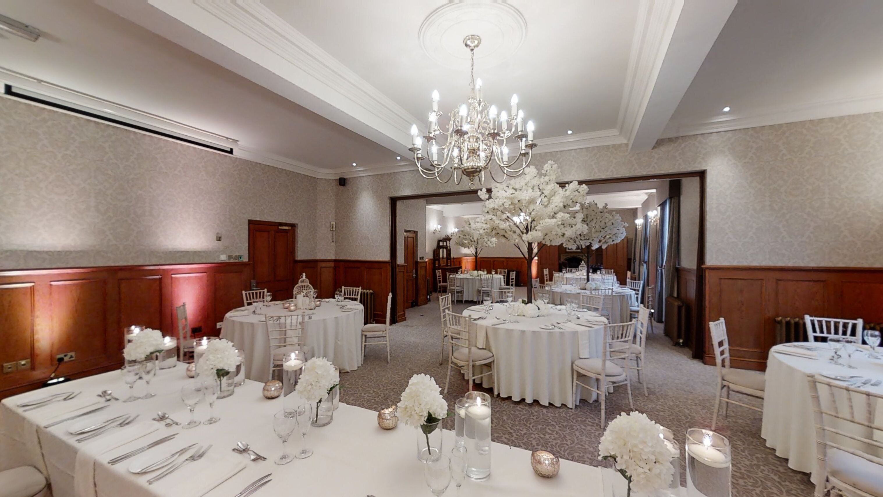 Een feestzaal met ronde tafels gedekt voor een diner, witte bloemstukken en een kroonluchter.