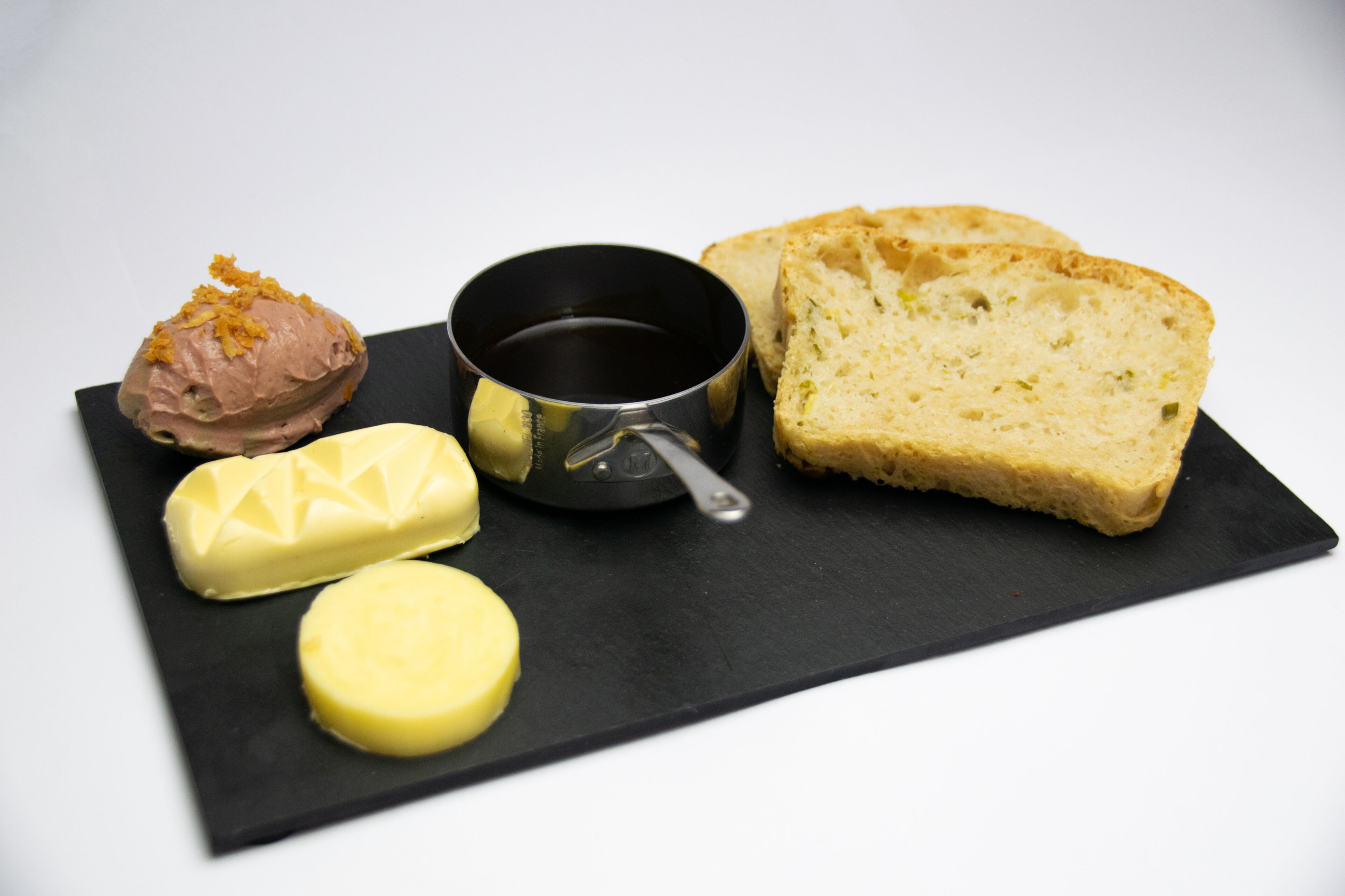 Una bandeja de pizarra negra con tres tipos de mantequilla, una pequeña vasija negra y dos rebanadas de pan.