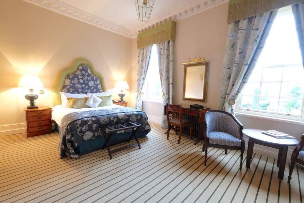 A hotel room with a bed, bedside lamps, a chair, a table, a mirror, and a window with curtains.
