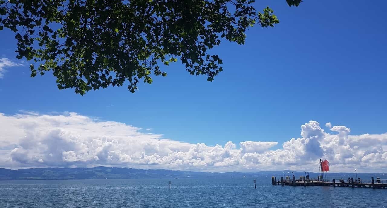 A clear view of a lake with a dock extending into the water, surrounded by mountains and a partly cloudy sky.