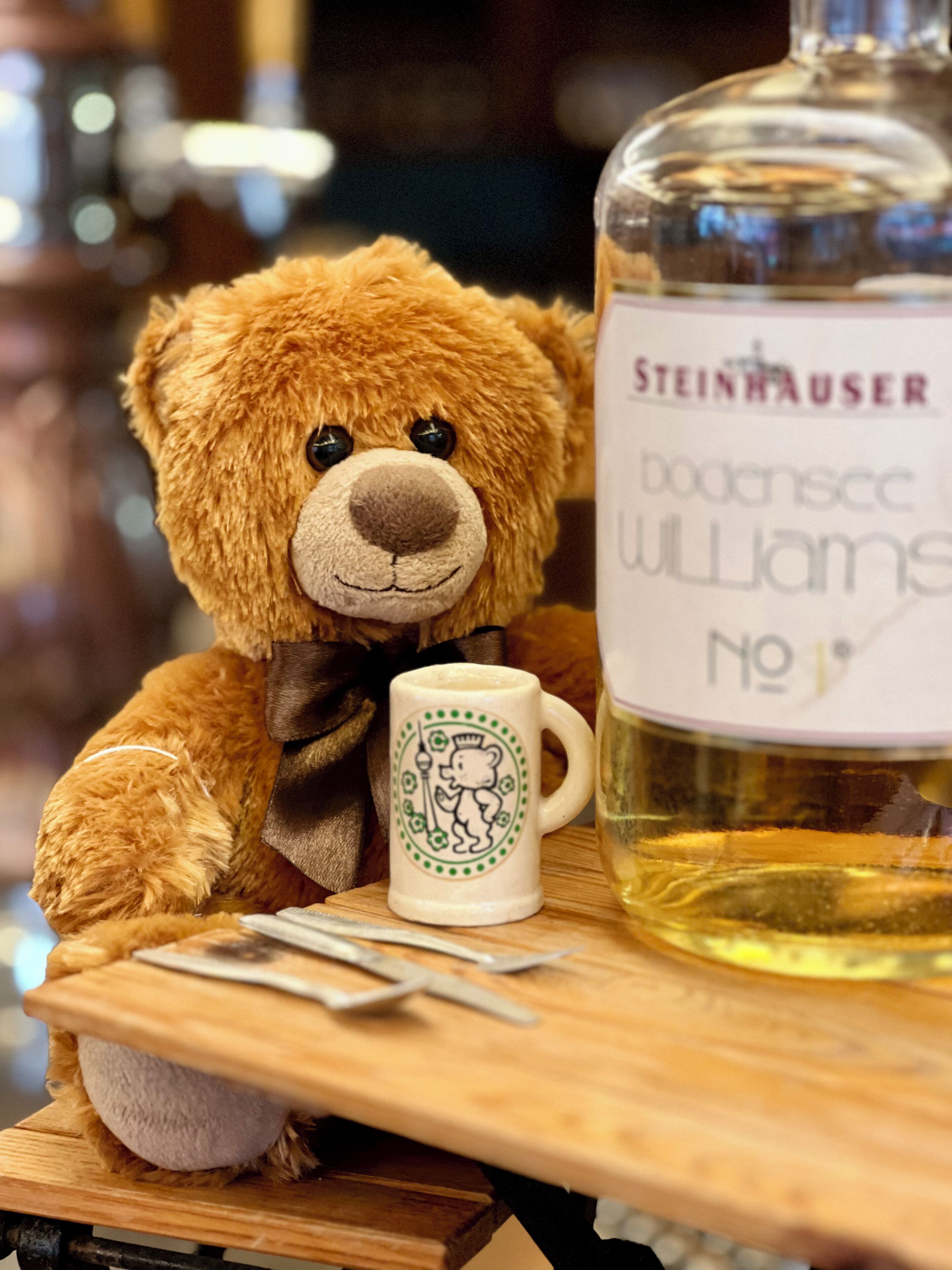 Een zachtaardig teddybeer, een fles Steinauer Donnase Williams No. 1, en een mok met een berendesign op een houten tafel.