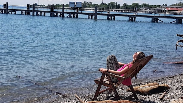 A person sitting on a wooden chair by a river, with a bridge and buildings in the background. - Teddybärenhotel (Kressbronn)