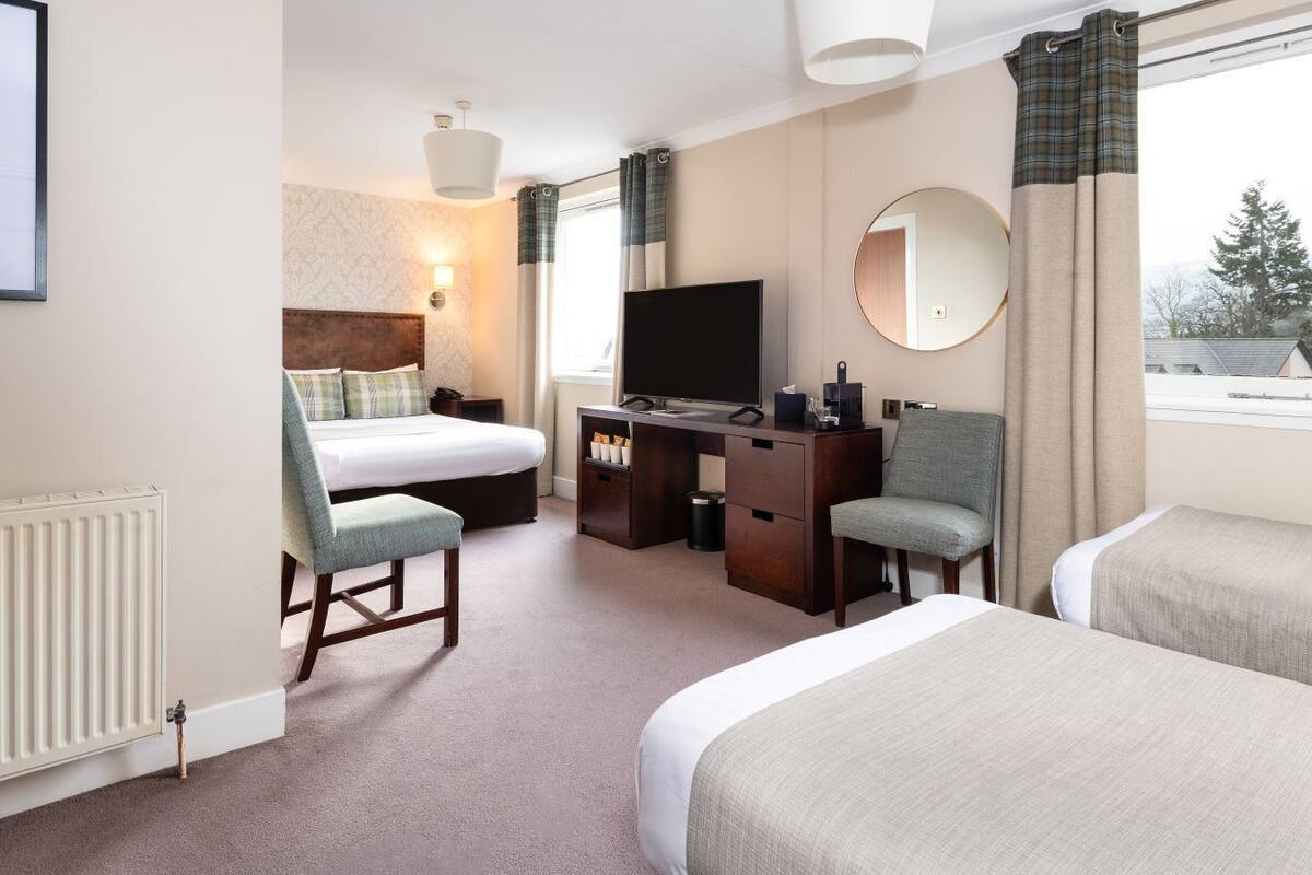 A hotel room with a bed, a desk, a chair, a TV, a dresser, and a window with curtains.