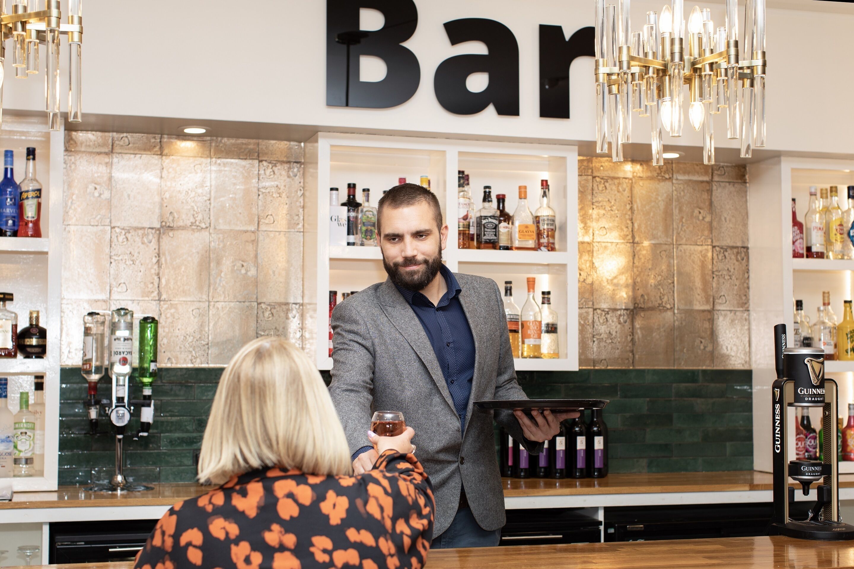 Uma área de bar com prateleiras de garrafas de bebidas, um bartender e um cliente segurando um copo.