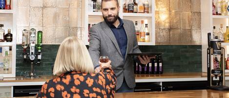 Uma área de bar com prateleiras de garrafas de bebidas, um bartender e um cliente segurando um copo.