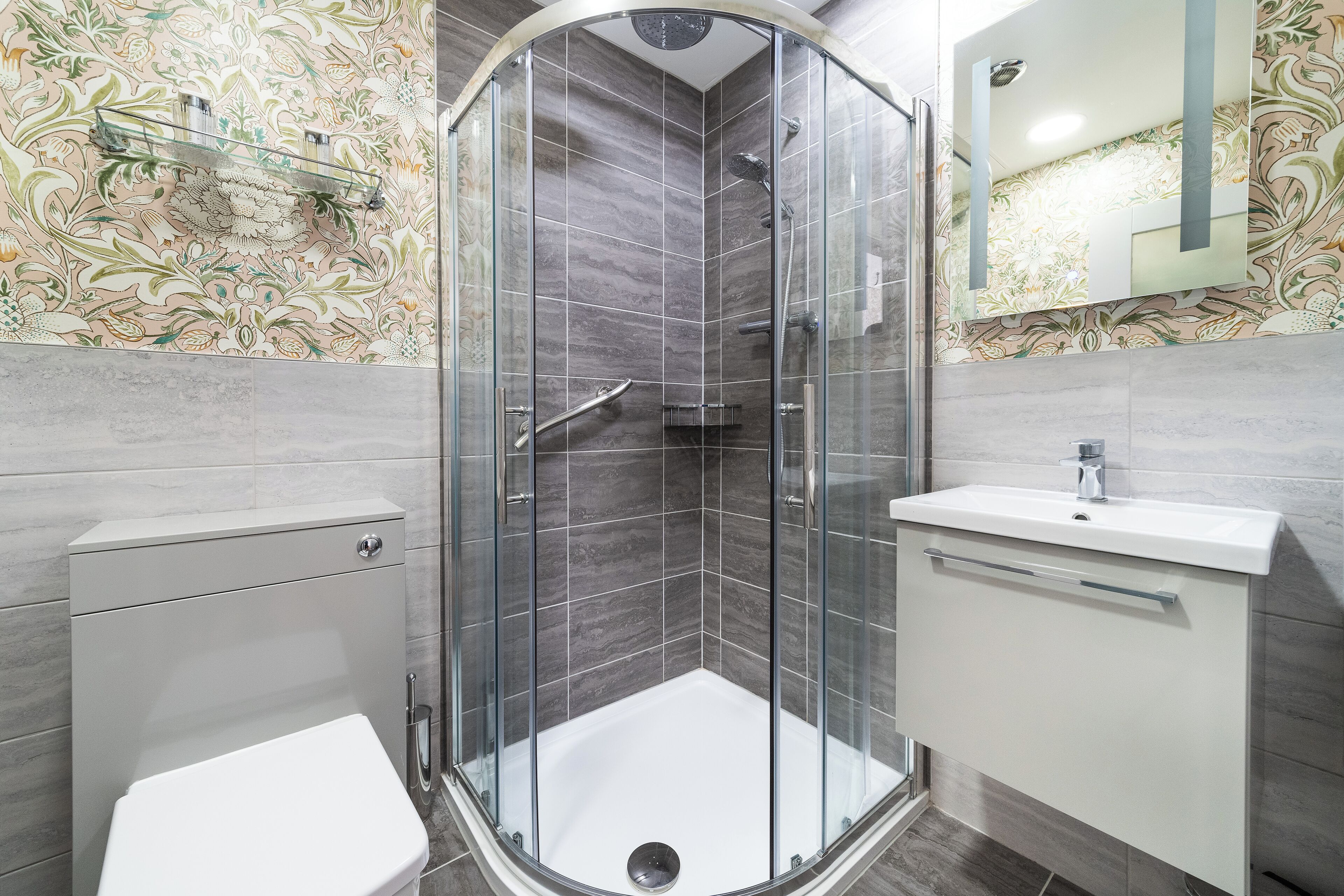 Et moderne badeværelse med en brusekabine i glas, et hvidt toilet og en vask med et spejl over.