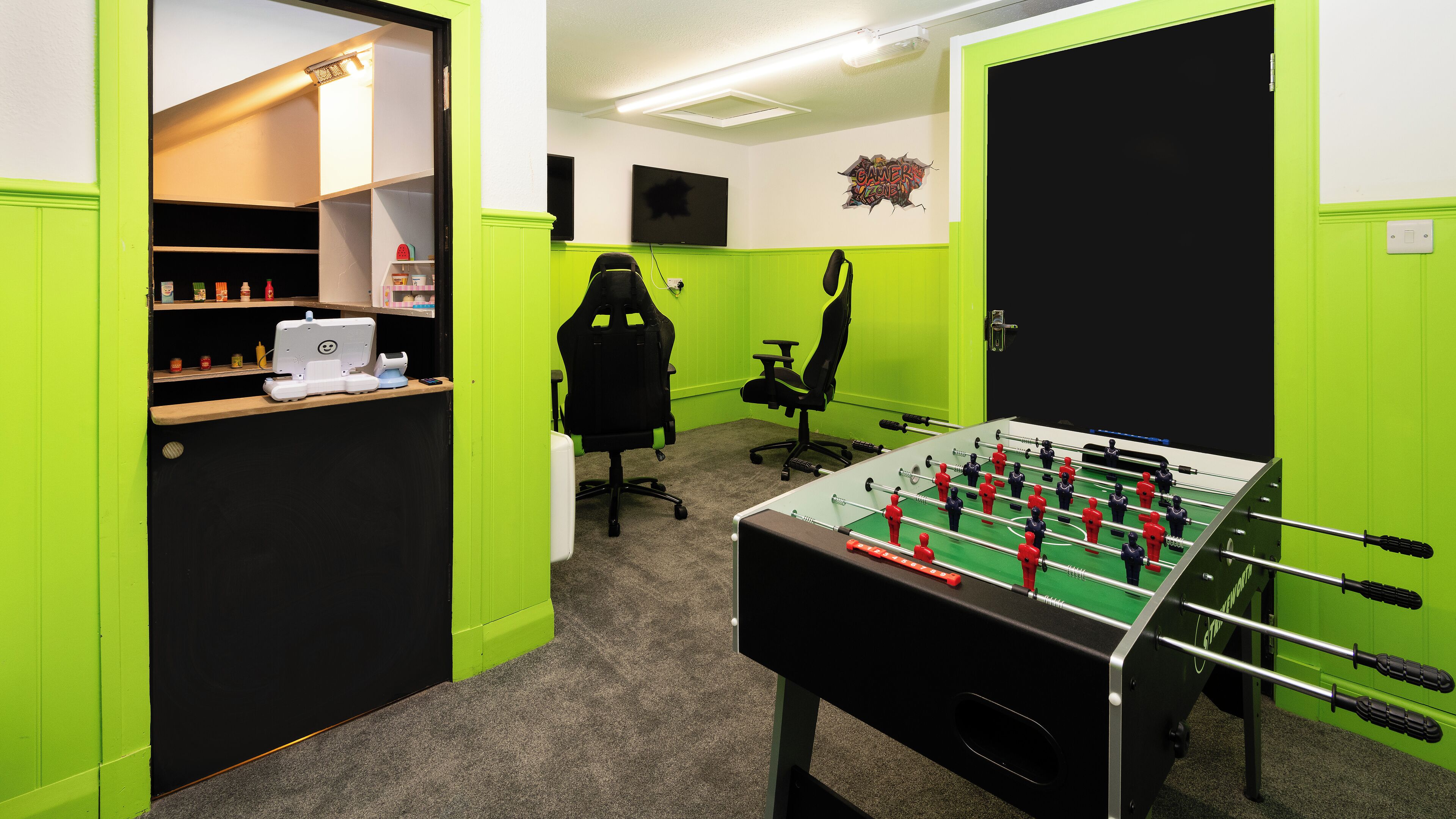 A room with a foosball table, two gaming chairs, a desk with a sewing machine, and a shelf with various items.