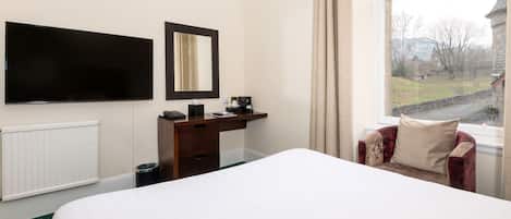A hotel room with a bed, a desk with a telephone and a coffee maker, a TV mounted on the wall, and a window with a view of a building and trees.