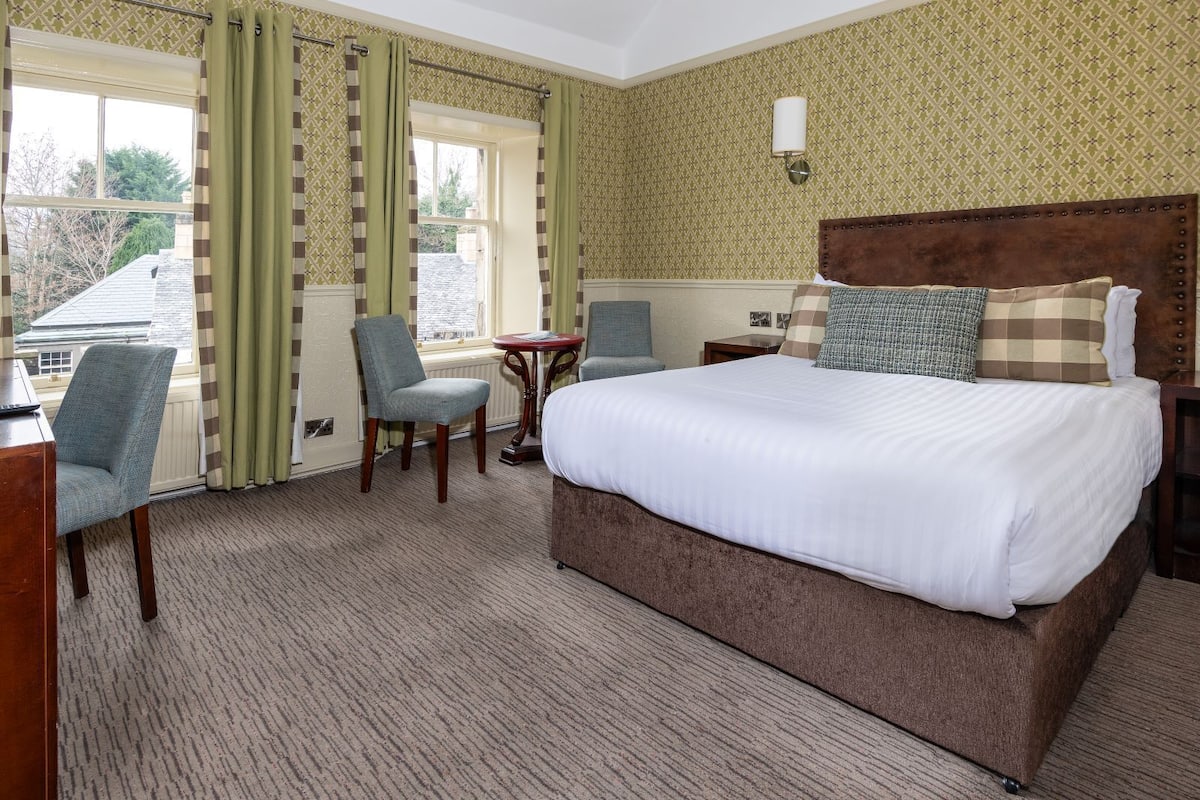 A hotel room with a large bed, a chair, a small table, and a window with curtains.