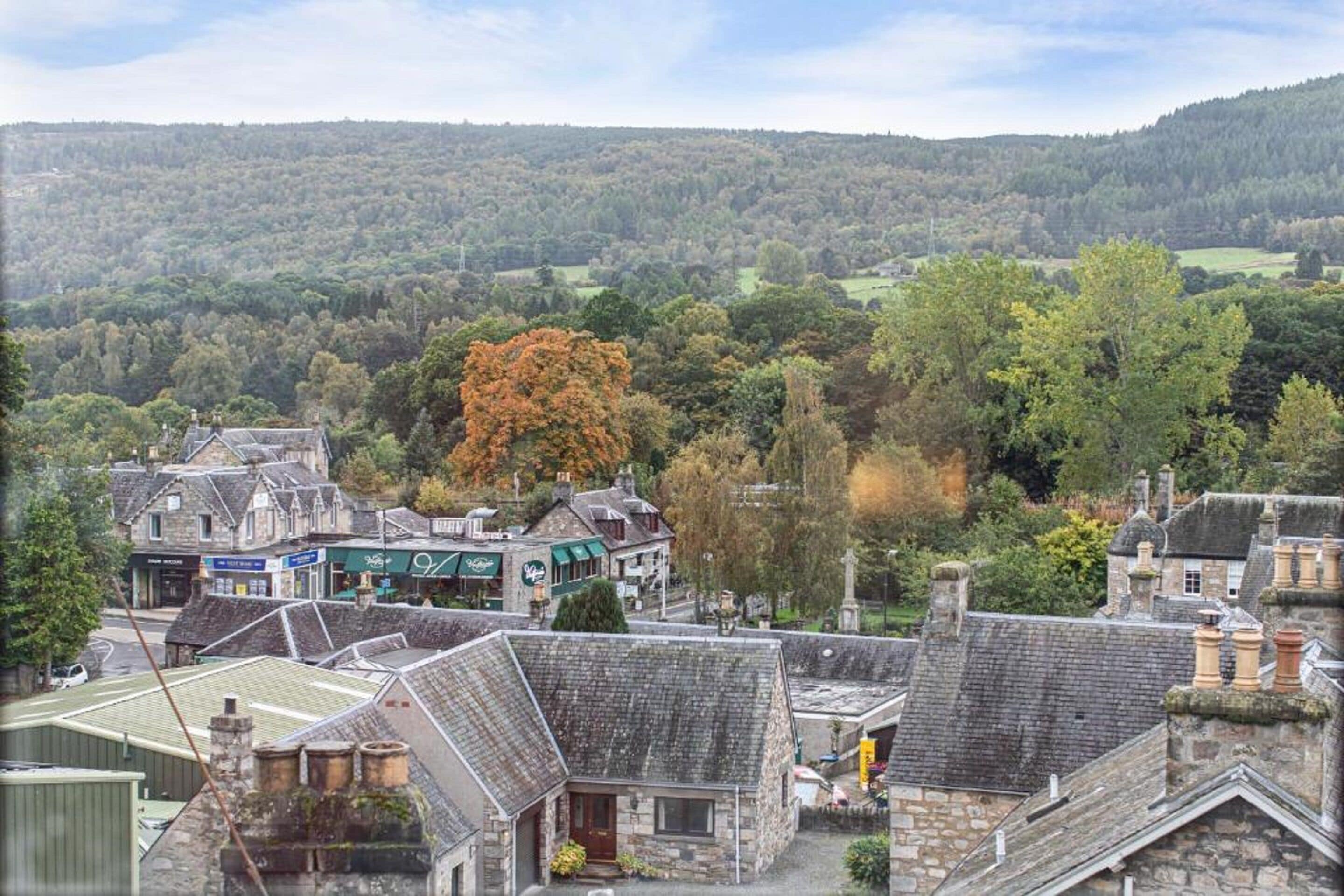 A traditional stone building with a steeply pitched roof, surrounded by a small town with visible shops and a forested hillside in the background.