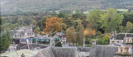 A traditional stone building with a steeply pitched roof, surrounded by a small town with visible shops and a forested hillside in the background.