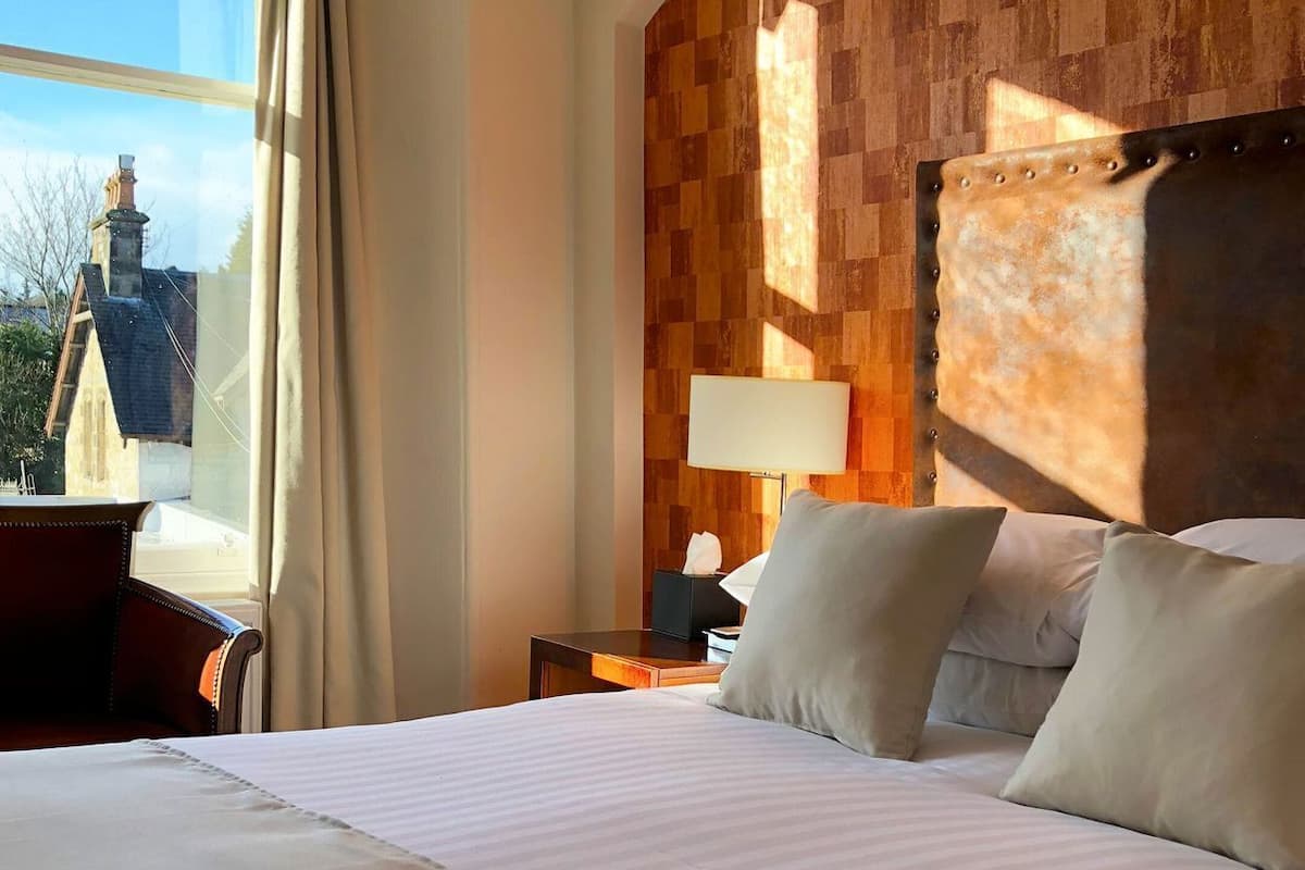 A hotel room with a large bed, a nightstand, a lamp, and a view of buildings outside.