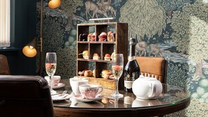 A dining table with a floral wallpaper backdrop, a wooden dessert display, and a wine bottle.