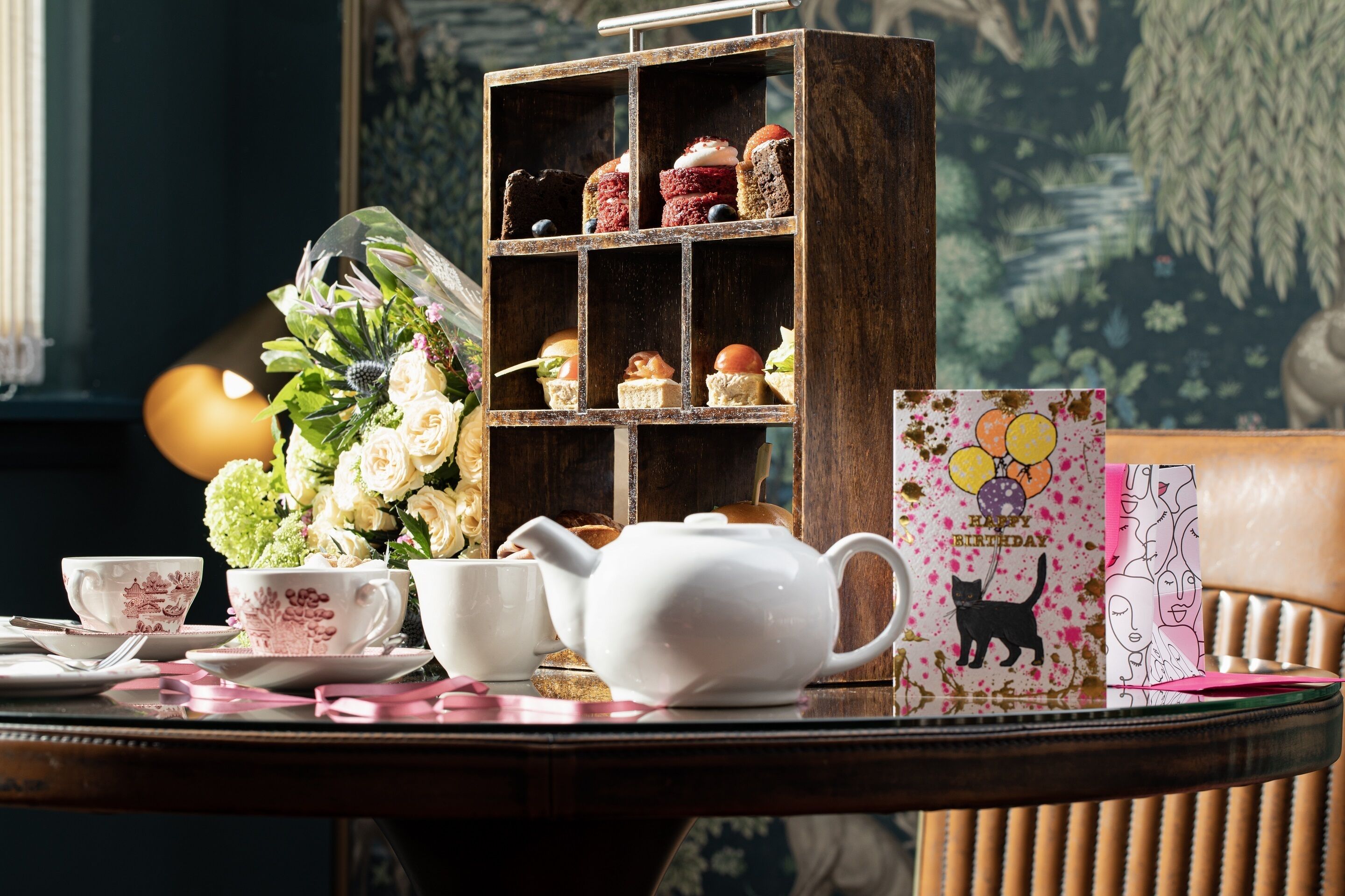 A table with a teapot, cups, a floral arrangement, a shelf with pastries, and a birthday card.
