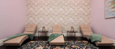 A room with a patterned wallpaper, a floral carpet, and two massage chairs with side tables.