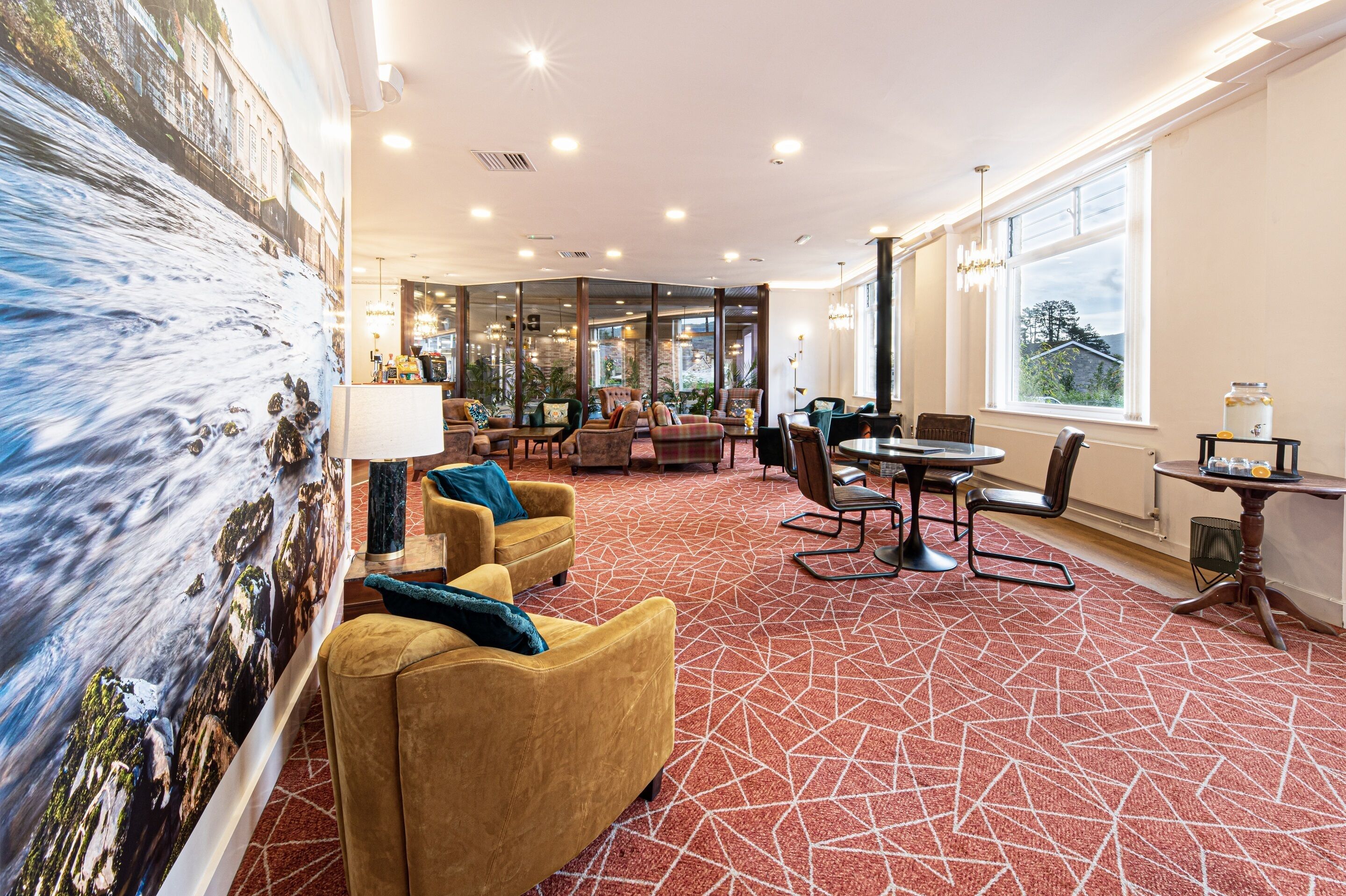 A hotel lobby with a red patterned carpet, yellow armchairs, a dining area with a round table, and a large mural of a river scene on the left wall.