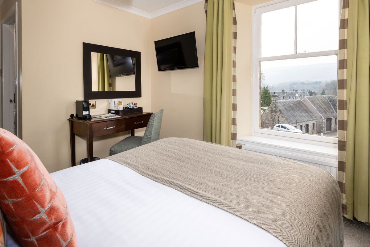 Habitación de hotel con cama, escritorio con computadora, televisor y ventana con vista a edificios.