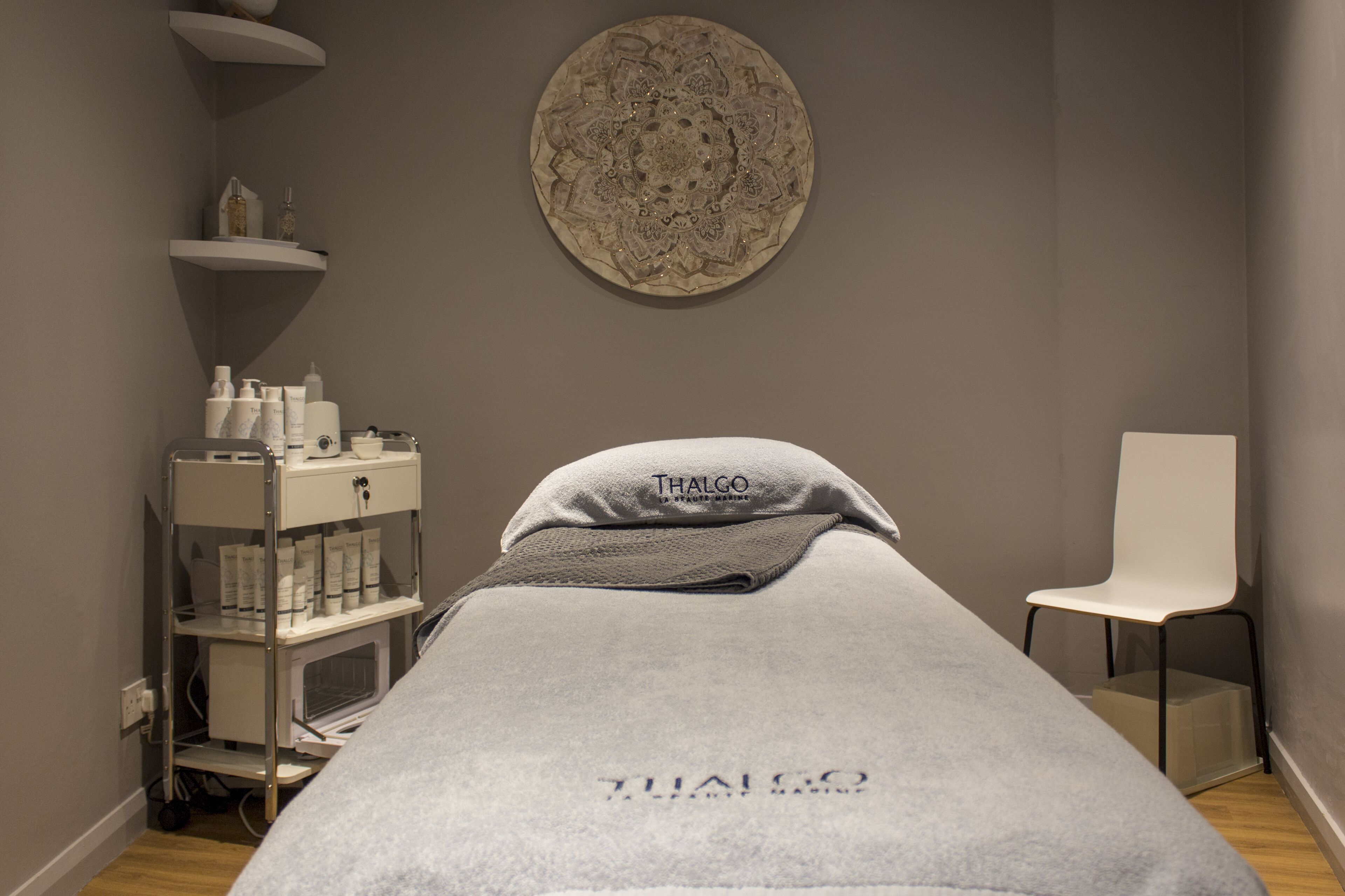 A treatment room with a massage table, a white chair, and a shelf with various bottles and containers.