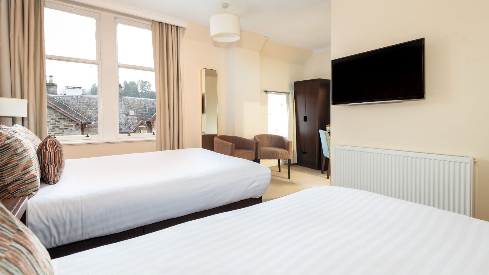 A hotel room with two beds, a TV, a chair, and a view of a stone building outside.
