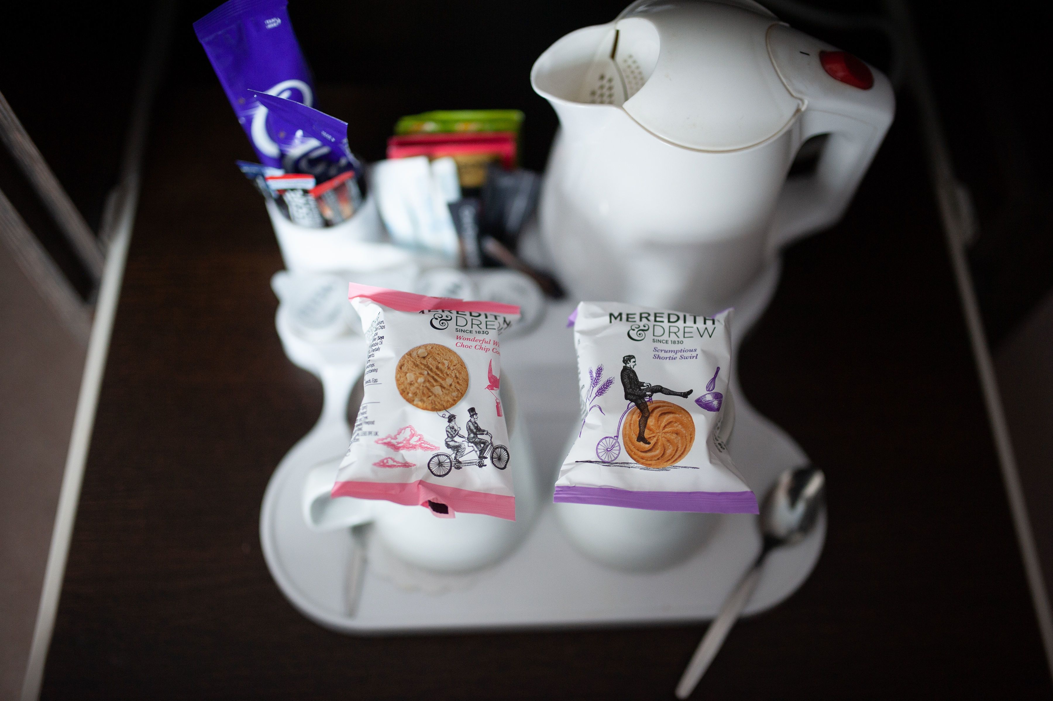Una máquina de café blanca con un botón rojo, dos paquetes de galletas Shortie Squirrel de Meredith Drew y un paquete púrpura de chocolate Cadbury Dairy Milk.