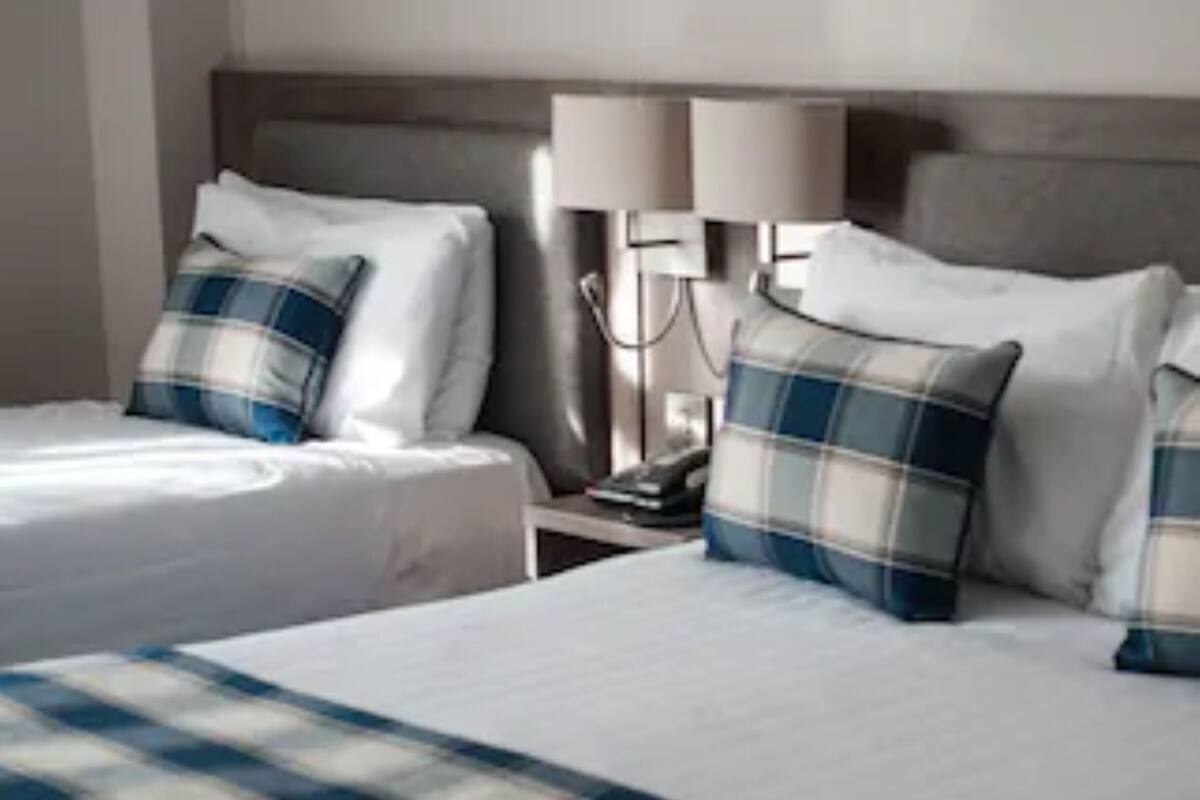 A hotel room with two beds, each with a plaid blanket and pillows. There is a nightstand with a lamp and a phone.