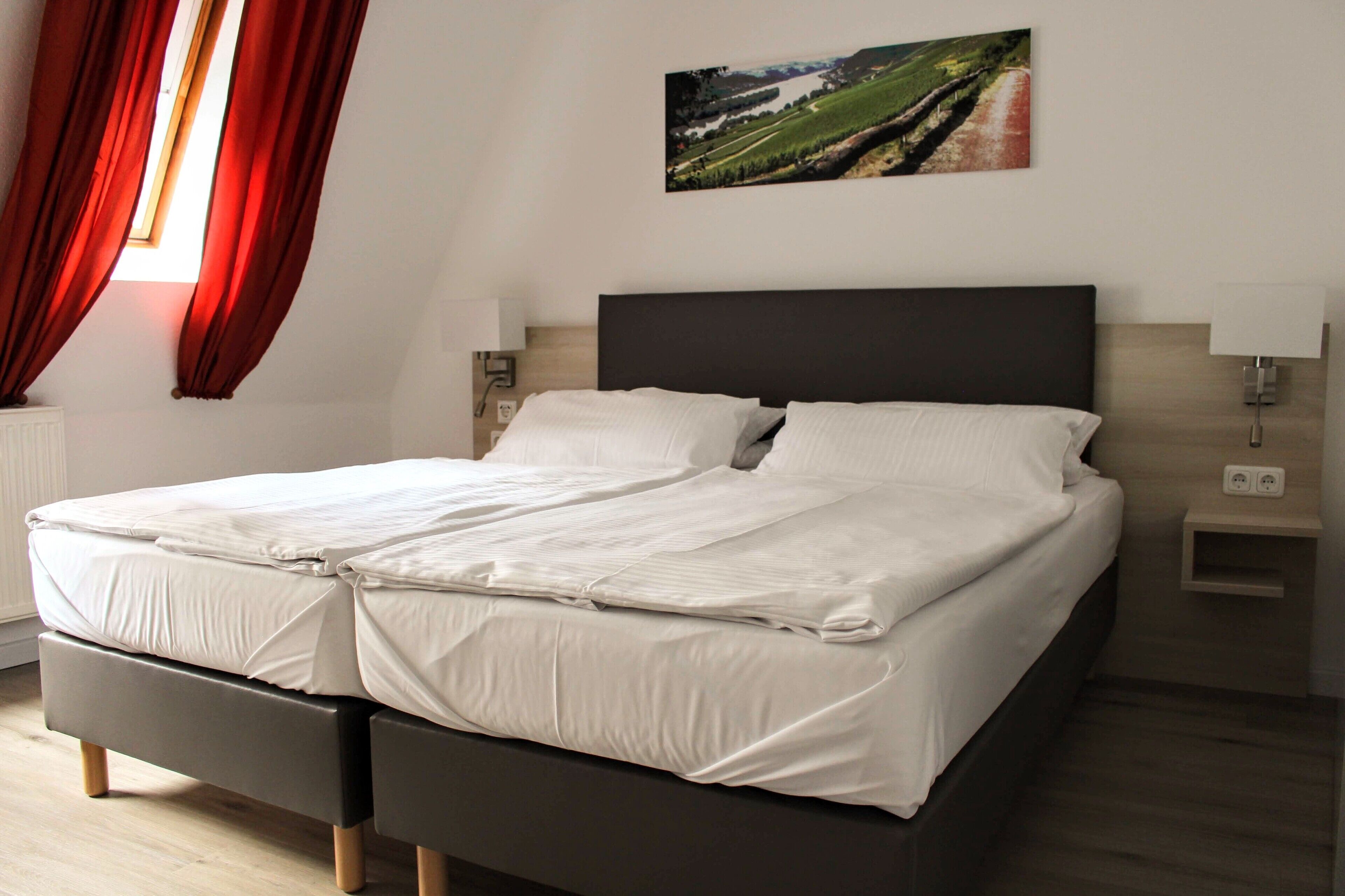 Uma cama de casal com lençóis brancos, cabeceira escura, duas mesas de cabeceira com abajures e um quadro com paisagem na parede.