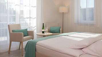 A hotel room with a bed, a chair, a small table with a bottle and glasses, and a lamp.