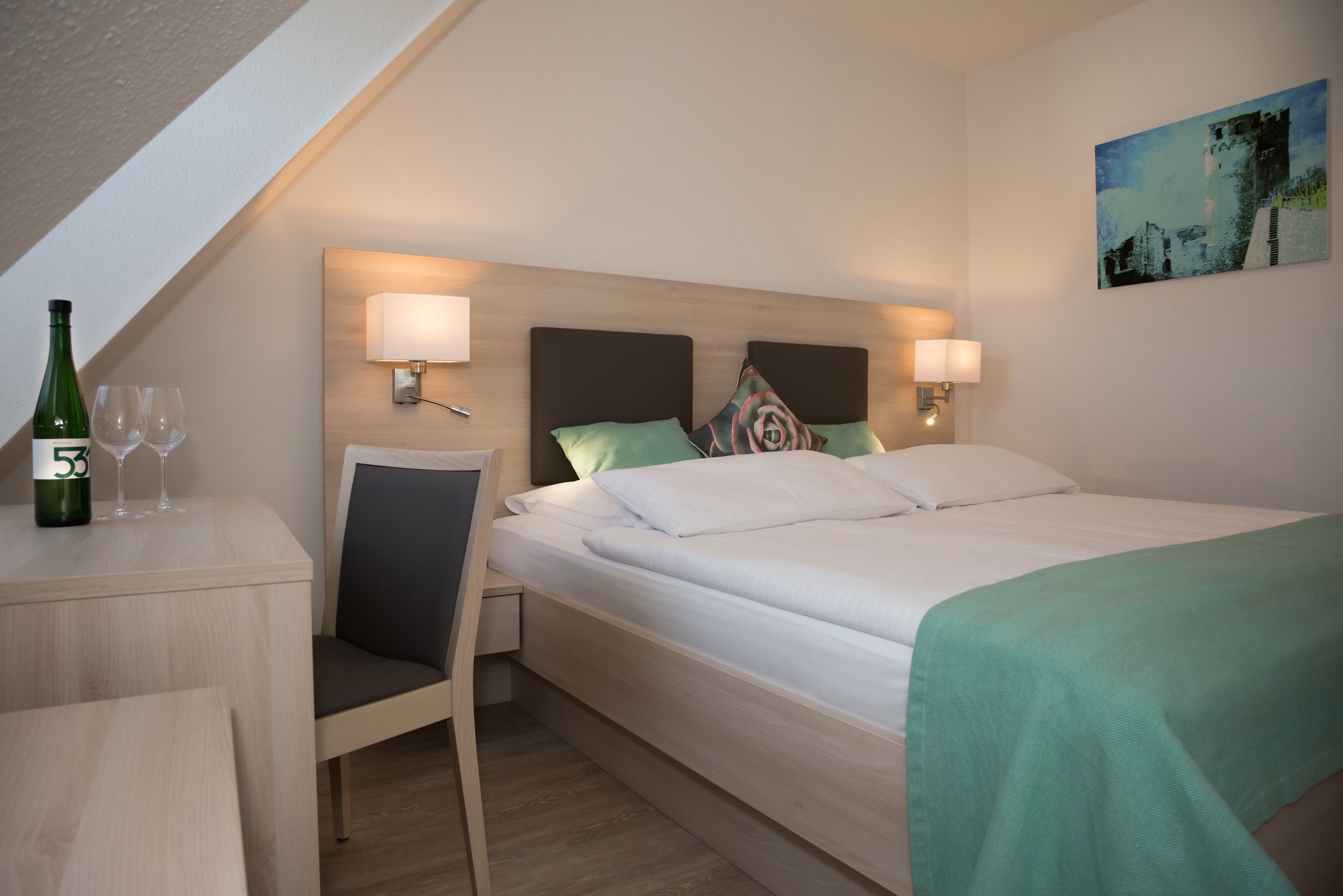 Habitación de hotel con cama, lámparas de noche, escritorio con botella y vasos, y un cuadro en la pared.