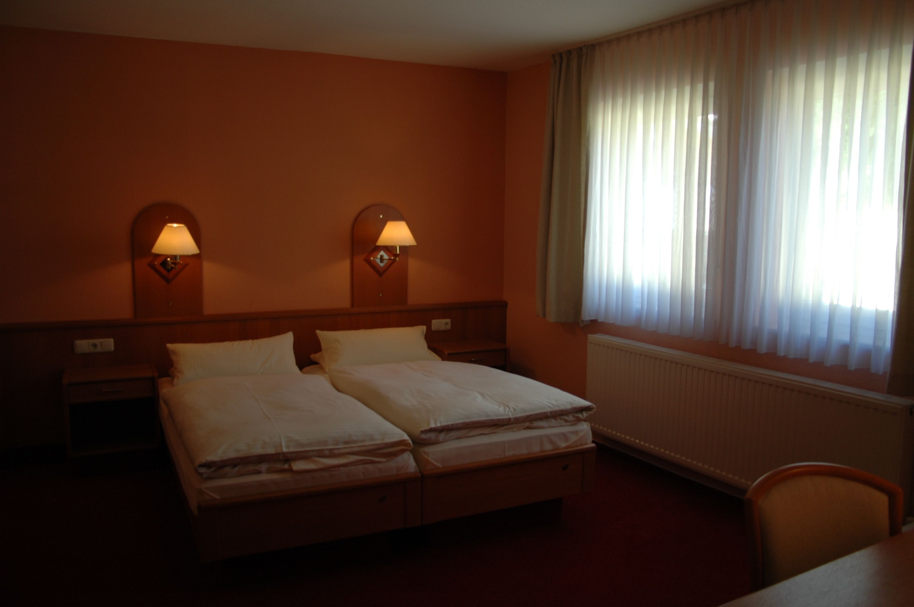 A hotel room with a double bed, two bedside tables, two wall-mounted lamps, a window with sheer curtains, and a radiator.