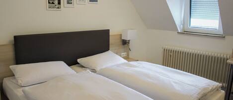 A twin bed room with a dark headboard, white bedding, a small bedside table with a lamp, and a window with blinds.
