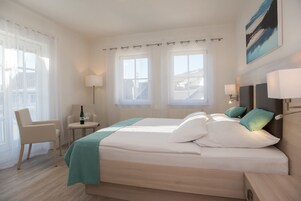 Una habitación de hotel moderna con una cama grande, dos lámparas de noche, una mesita redonda con una botella y un vaso, una silla y ventanas amplias con cortinas.