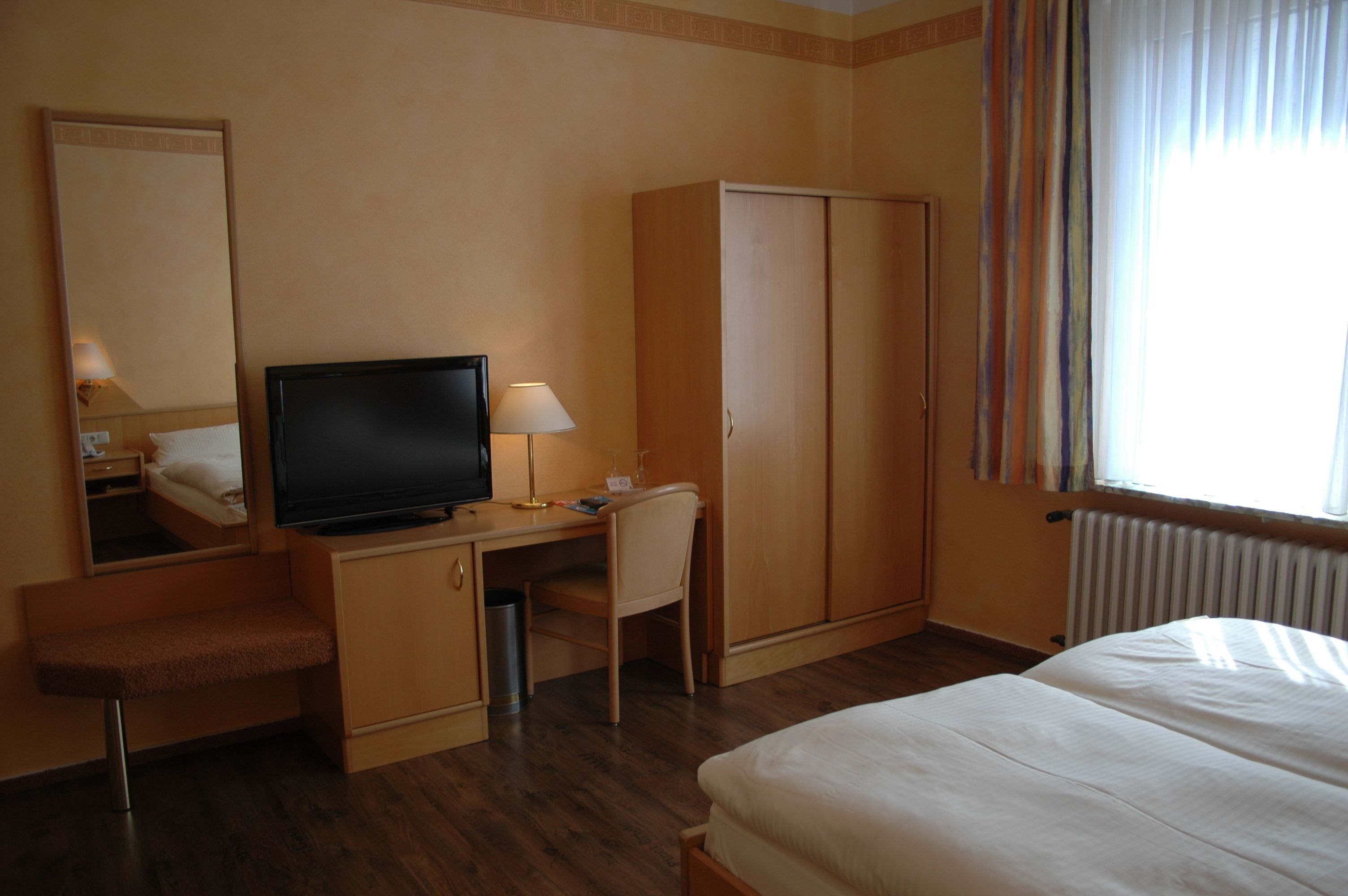 Ein Hotelzimmer mit einem Bett, einem Schreibtisch mit Fernseher, einem Sessel, einem Kleiderschrank und einem Fenster mit Vorhängen.
